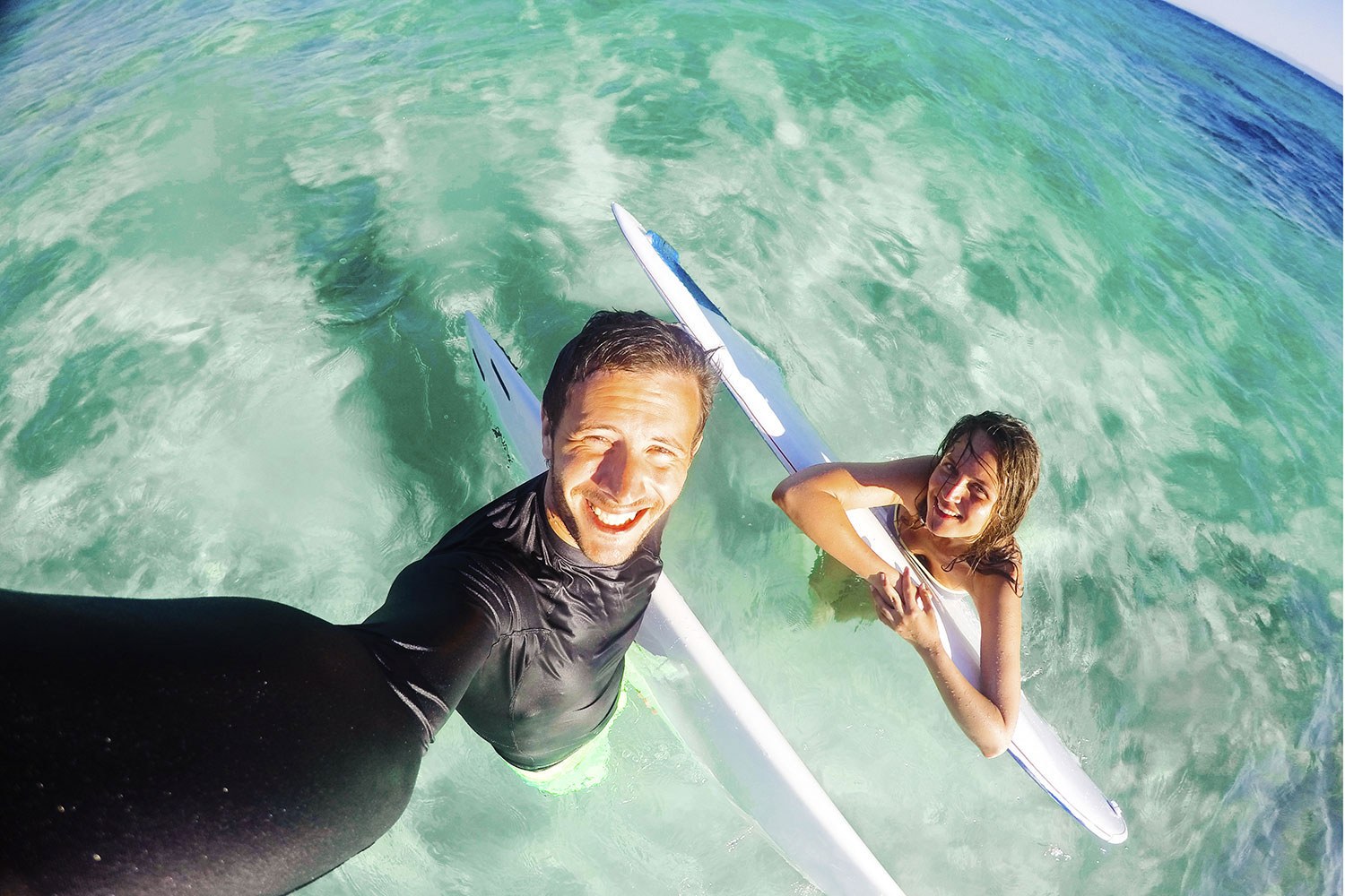 Ein Mann und eine Frau lächeln in türkisblauem Wasser und halten Surfbretter. Der Mann macht ein Selfie. Der Himmel ist sichtbar.