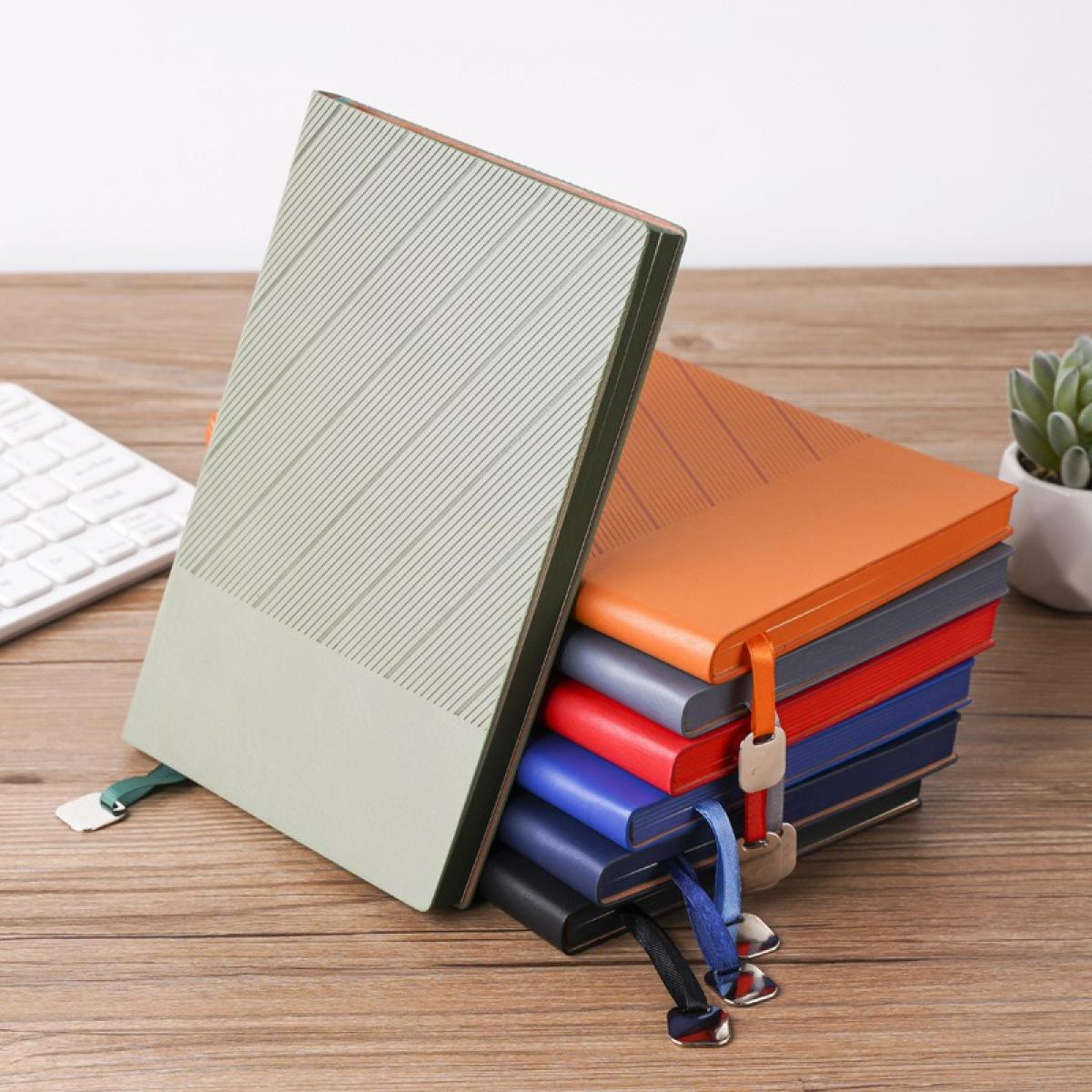 Ein Stapel bunter Notizbücher mit Lesezeichen, eines lehnt an dem Stapel. Eine Tastatur und eine Pflanze sind im Hintergrund auf einer Holzoberfläche.