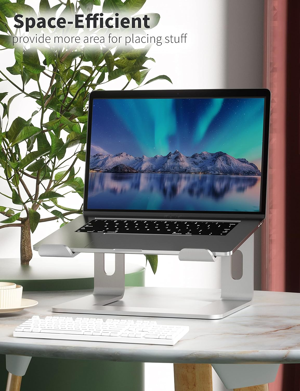 Laptop auf einem silbernen Ständer. Der Bildschirm zeigt Berge und Wasser. Eine weiße Tastatur liegt darunter auf einem Tisch.