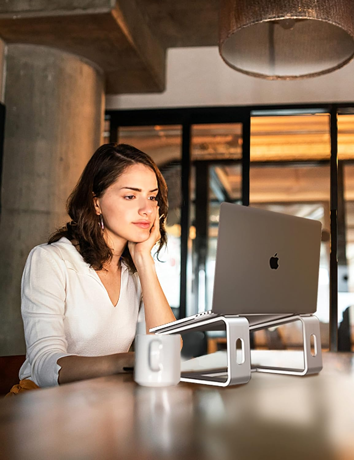 Eine Frau sitzt an einem Tisch und betrachtet einen Laptop auf einem Ständer. Neben dem Ständer steht eine weiße Tasse. Der Hintergrund zeigt ein verschwommenes Interieur.