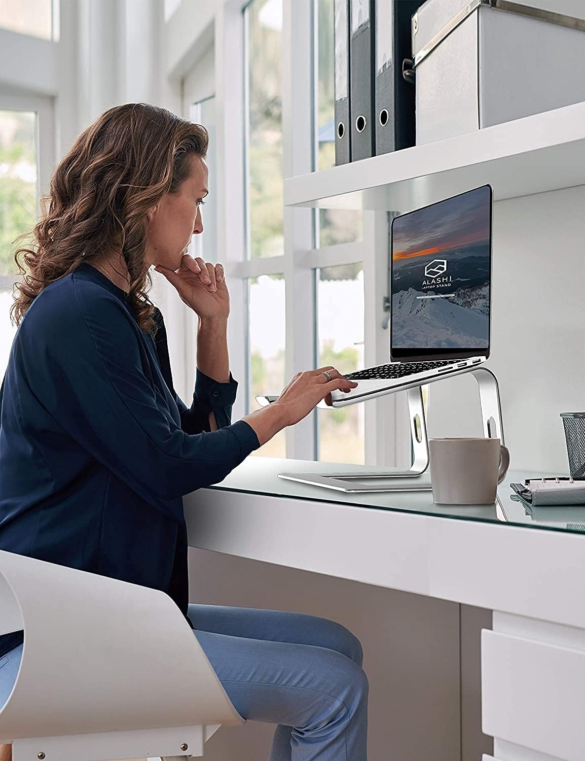 Frau benutzt Laptop am Schreibtisch. Eine Tasse und ein Laptopständer stehen auf dem Tisch. Regale und ein Fenster sind im Hintergrund.