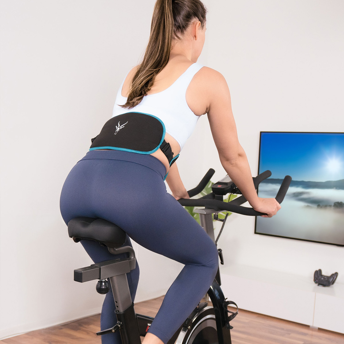 Eine Frau trainiert auf einem Heimtrainer. Sie trägt einen Stützgürtel und Leggings. Ein Fernseher zeigt eine Landschaft. Weiße Wände und ein weißer Schrank.