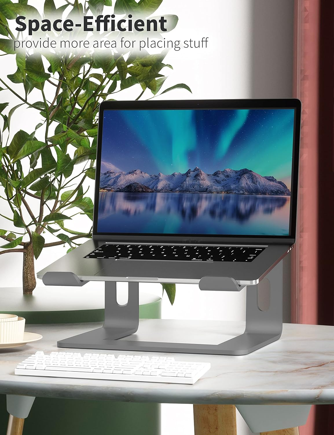 Laptop auf grauem Ständer, Tastatur davor, Hintergrund mit Pflanze und Berglandschaft auf dem Laptop-Bildschirm.