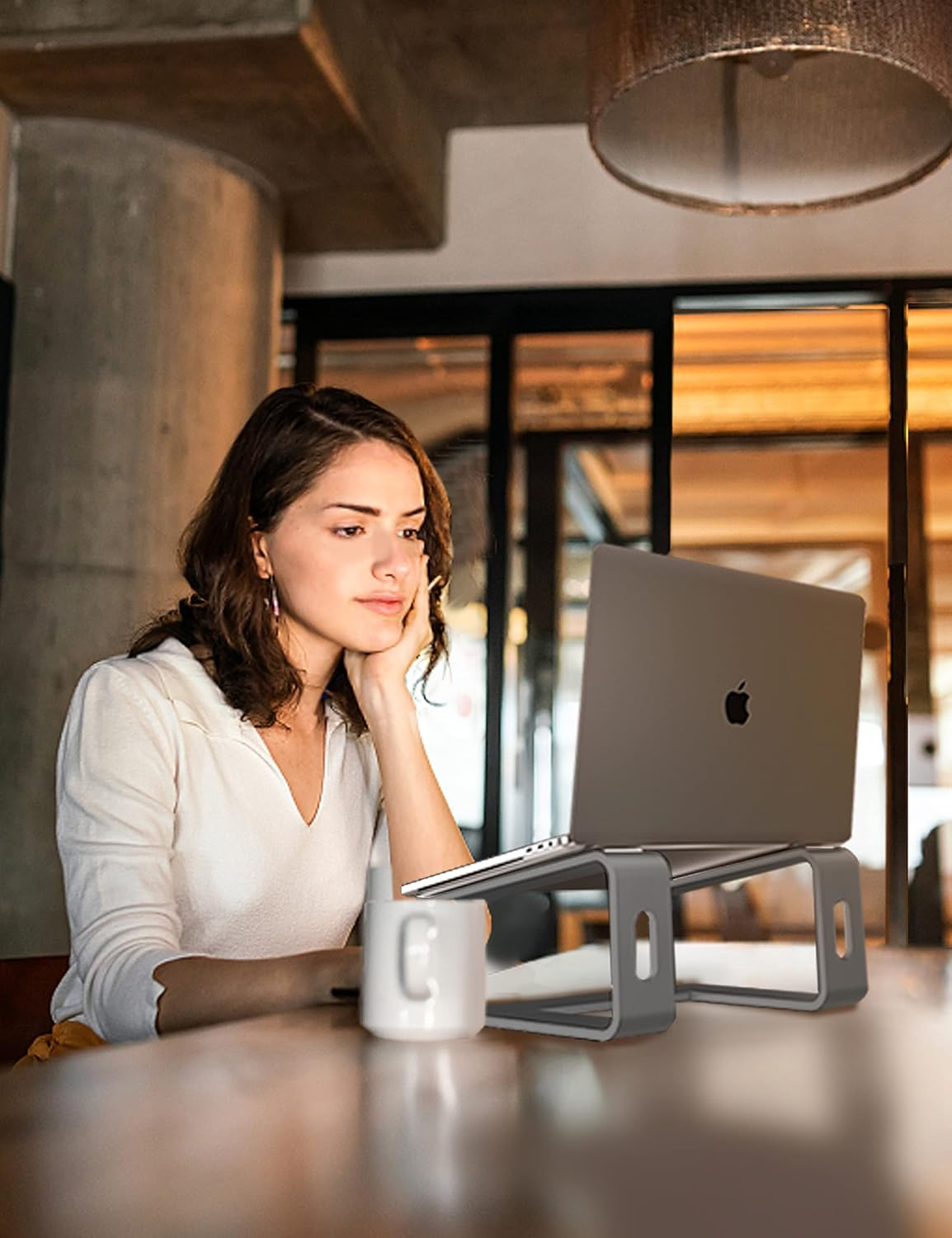 Eine Frau sitzt an einem Tisch und betrachtet einen Laptop. Eine Tasse und ein Laptopständer stehen auf dem Tisch. Der Hintergrund hat ein warmes Licht.