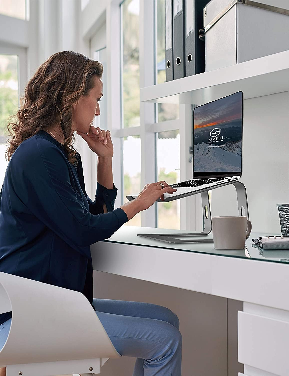 Frau arbeitet im hellen Büro an einem Laptop. Laptop steht auf einem Ständer. Becher und Akten sind sichtbar. Sie trägt Blau.