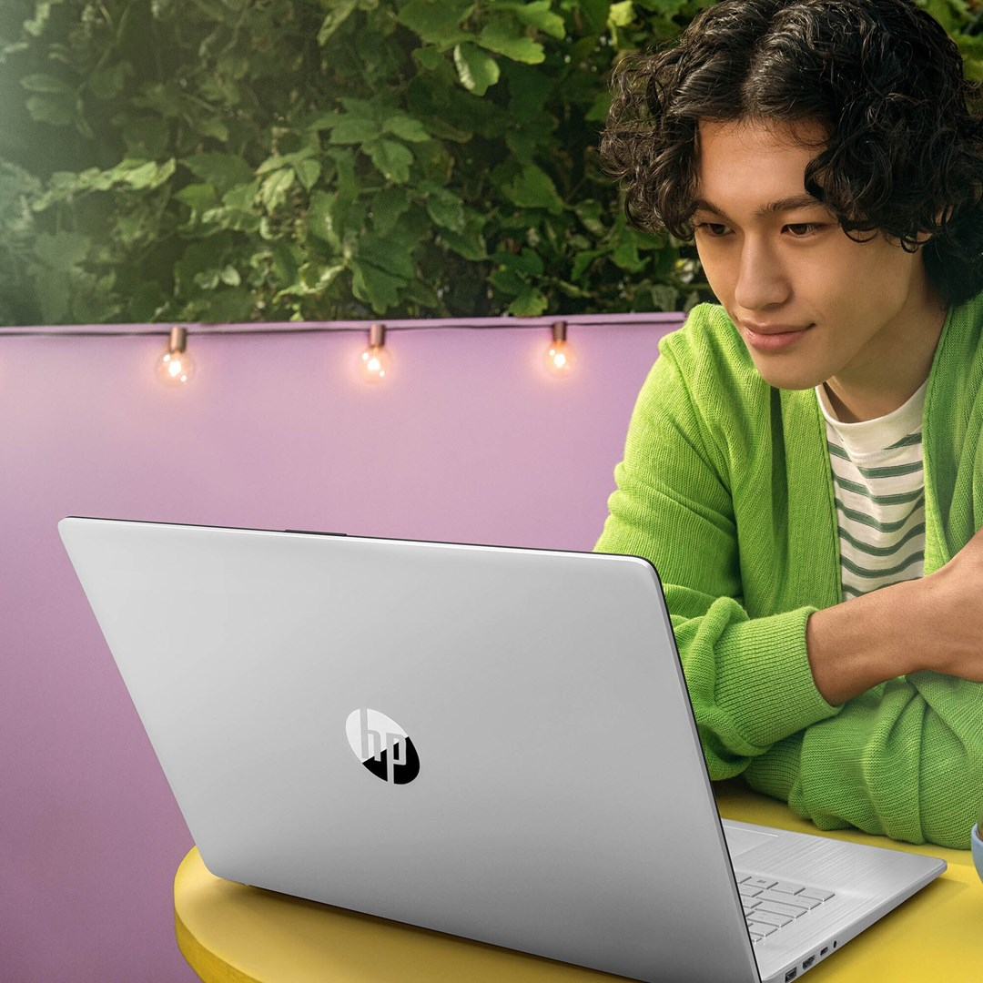 Młody mężczyzna w zielonym kardiganie patrzy na laptopa. Siedzi przy żółtym stole przed fioletową ścianą i zielonymi roślinami.