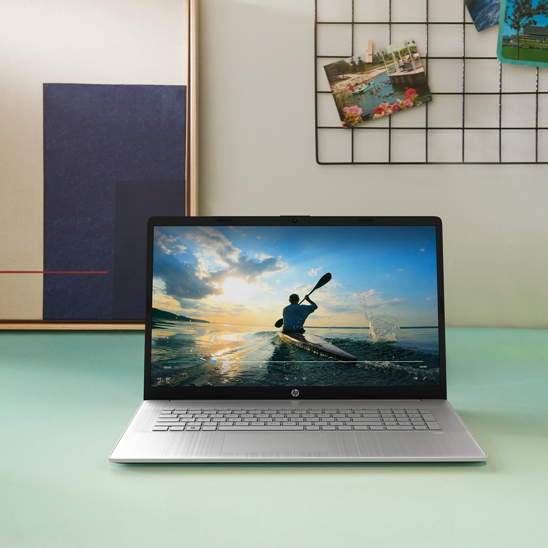 Laptop wyświetlający kajakarza na wodzie o zachodzie słońca. Laptop stoi na jasnozielonej powierzchni z grafiką na ścianie.