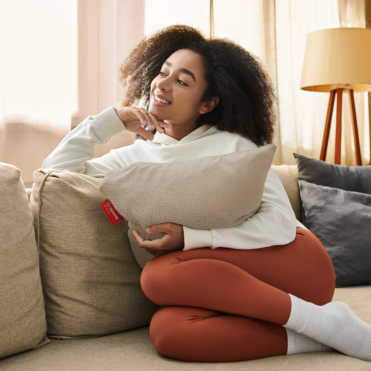 Young woman smiles, holding a pillow on a sofa. She wears a white hoodie, orange leggings, and white socks. A lamp is in the background.