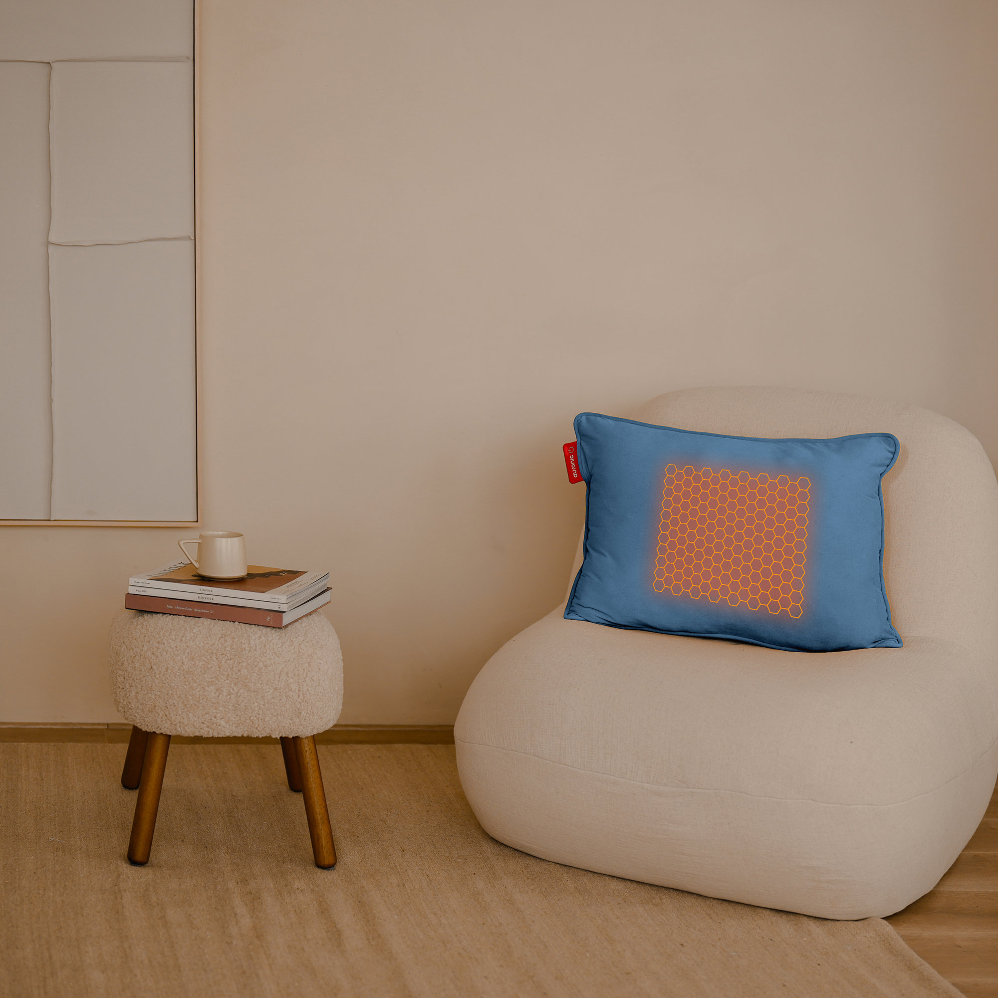 Een blauw kussen met een gloeiend oranje honingraatpatroon ligt op een witte stoel. Een kleine kruk heeft boeken en een mok.