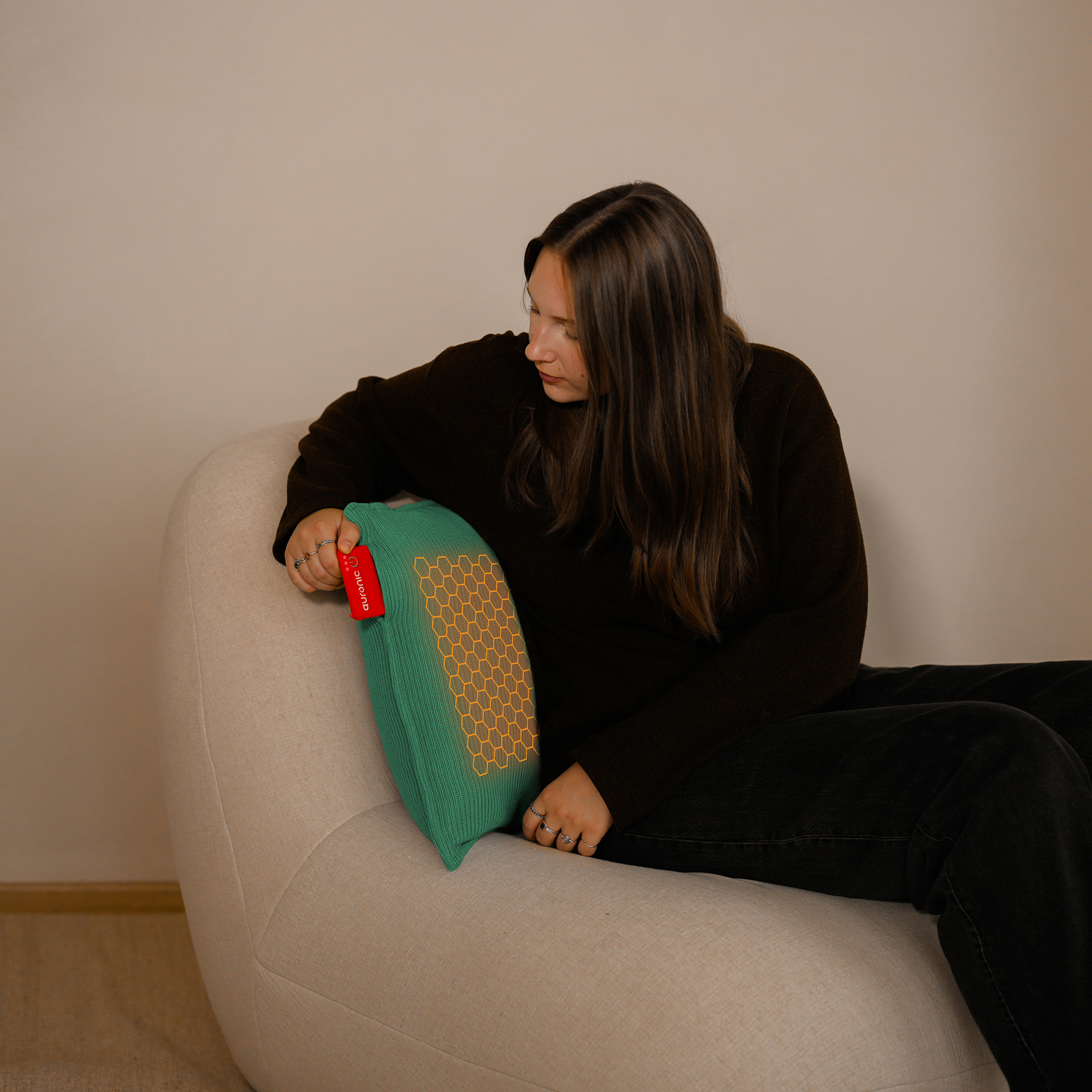 Femme assise dans un fauteuil beige tenant un coussin vert à motifs nid d'abeilles, portant des vêtements sombres.