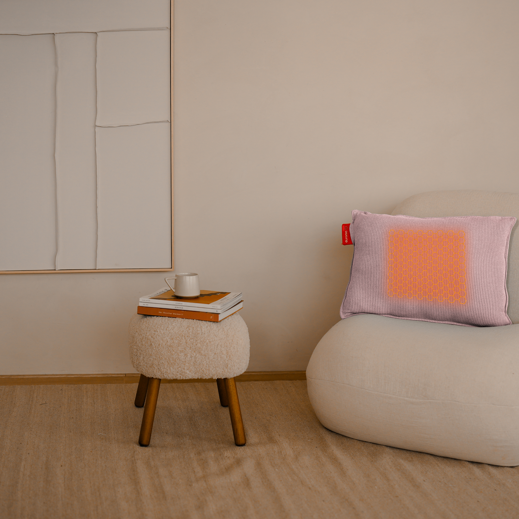 A cozy interior with a pink pillow with an orange center on a white chair. A stool with books and a mug is nearby.
