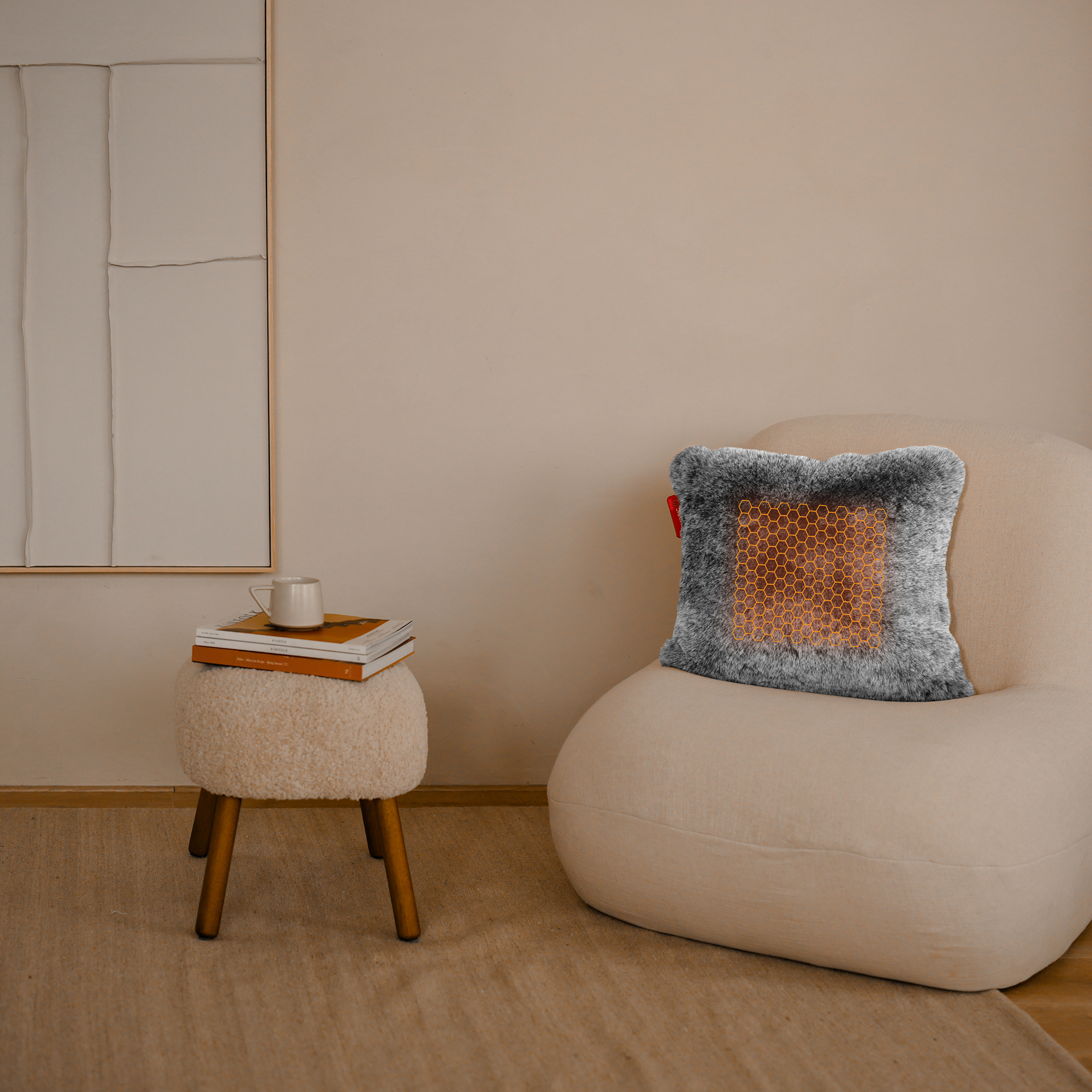 A cream chair with a grey pillow with a heating pattern. A small table with books and a cup. Beige wall.