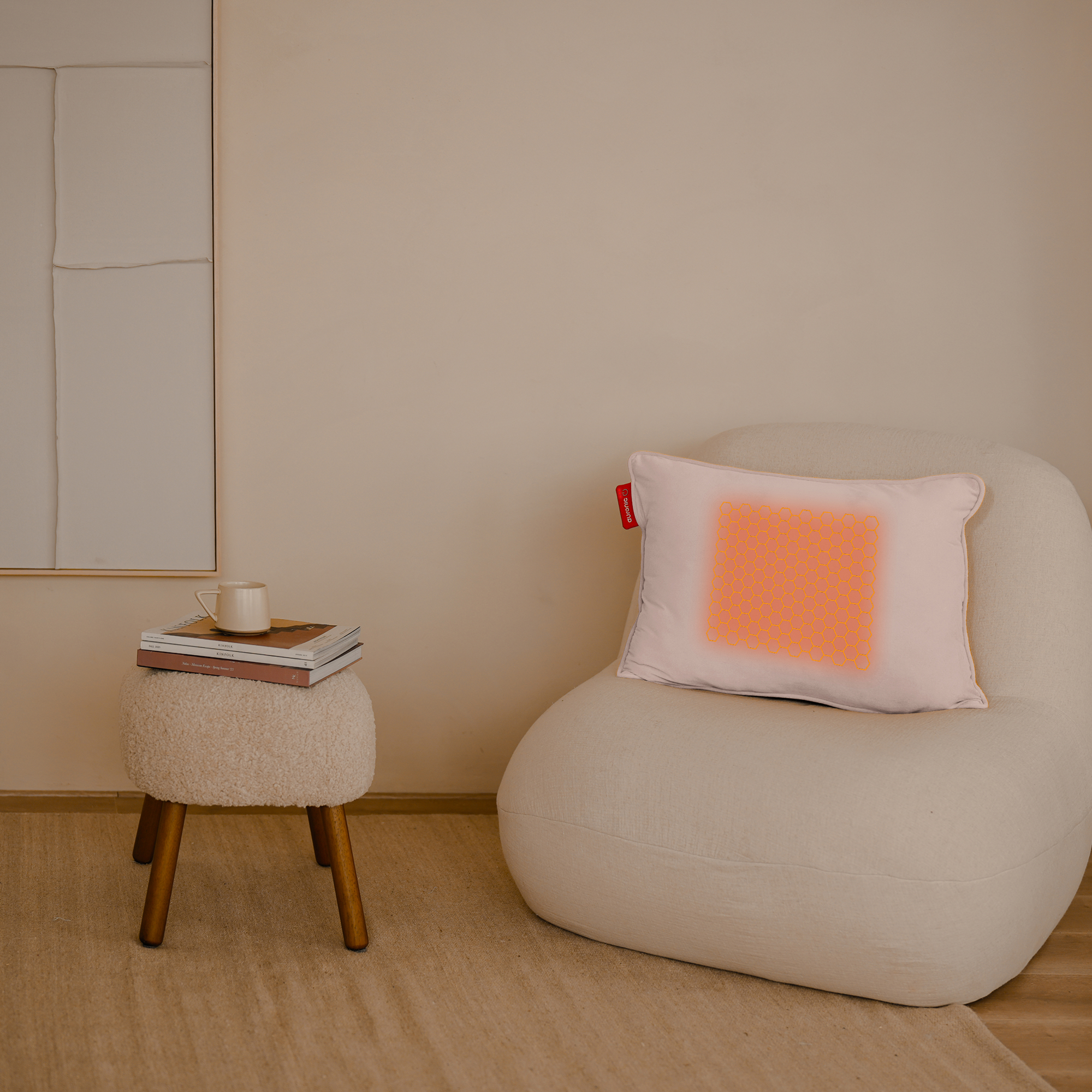 Een beige kamer met een stoel, boeken en een kruk. Een kussen met een oranje patroon ligt op de stoel.