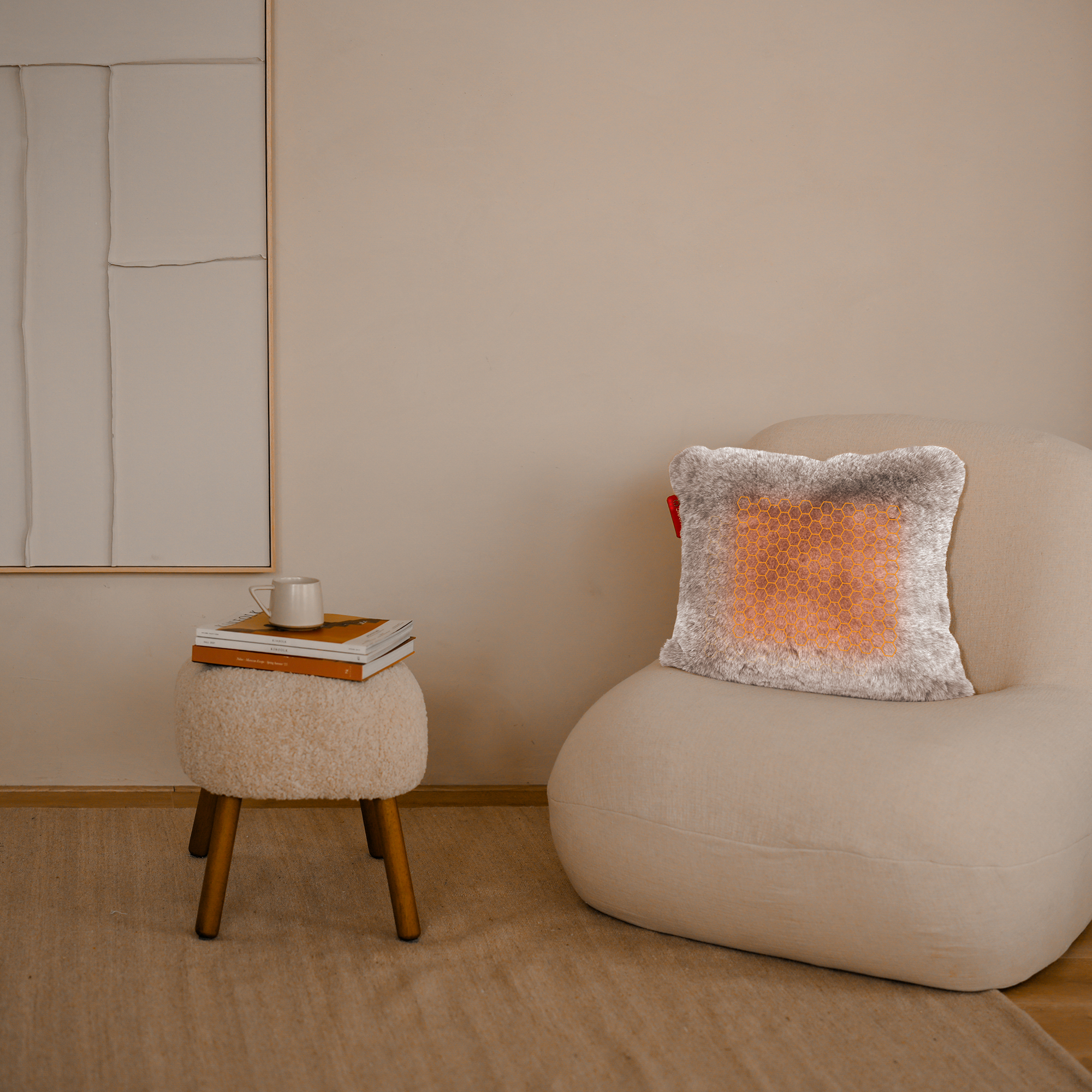 A beige room with a chair, a stool with books and a mug, and a decorative pillow with an orange pattern.