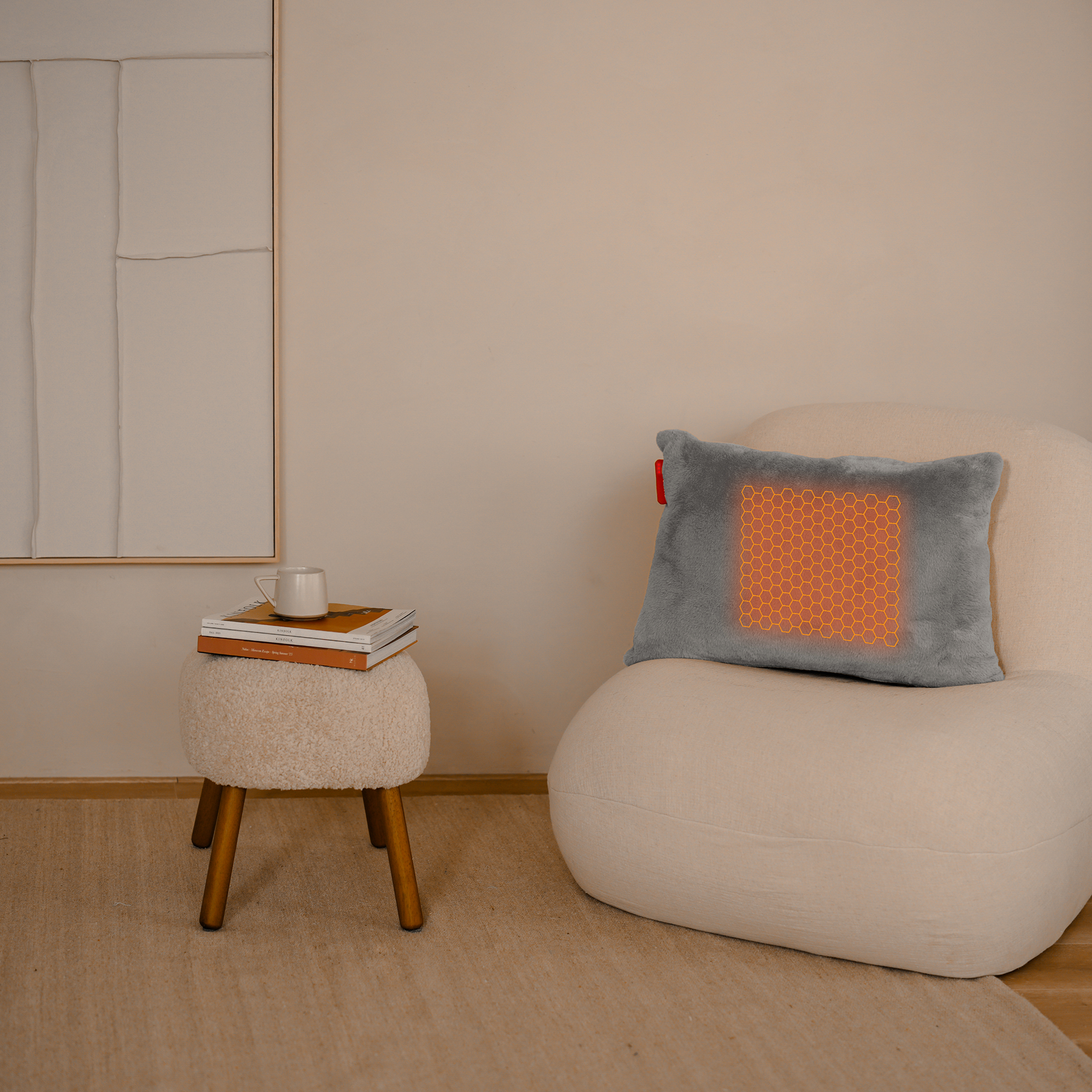 Een grijs kussen met een oranje verwarmingspatroon, een witte stoel, een krukje met boeken en een kopje, en een beige muur op de achtergrond.