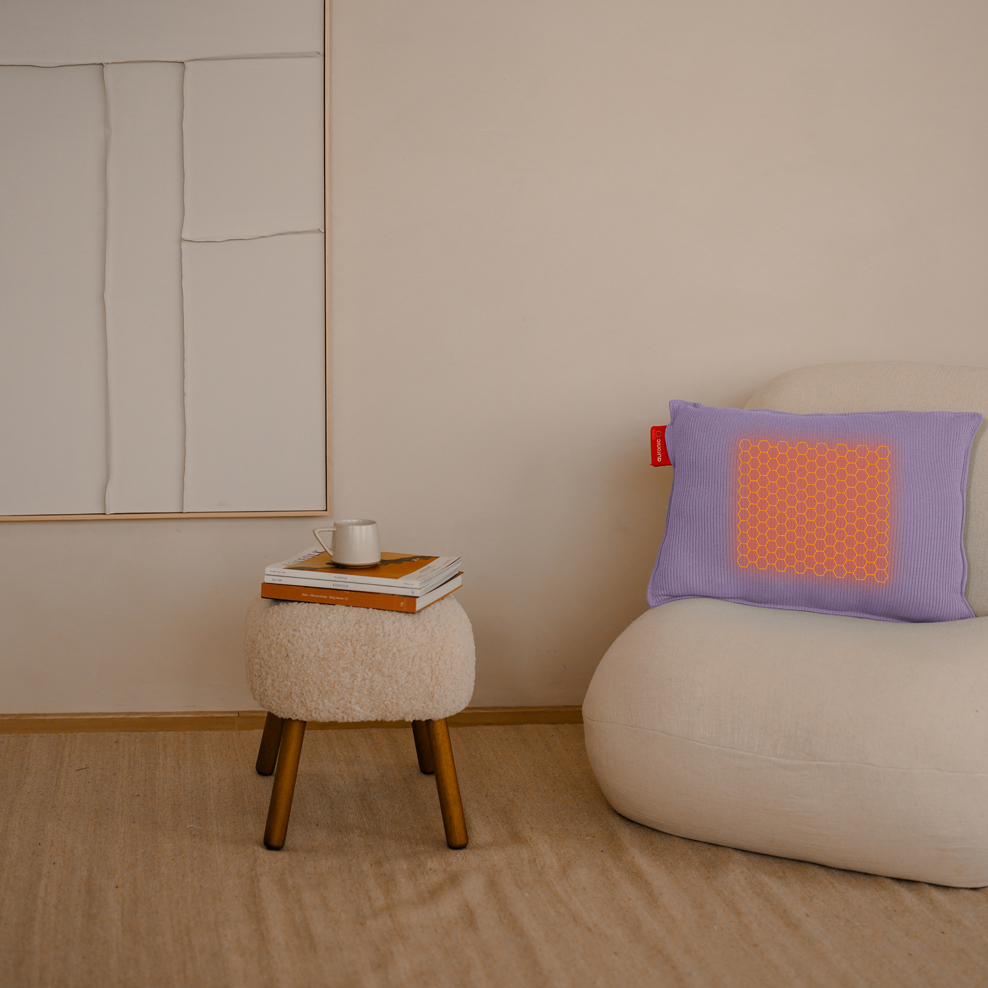Een kamer met een witte stoel, een kruk met boeken en een mok, en een paars kussen. Beige muren en schilderij.