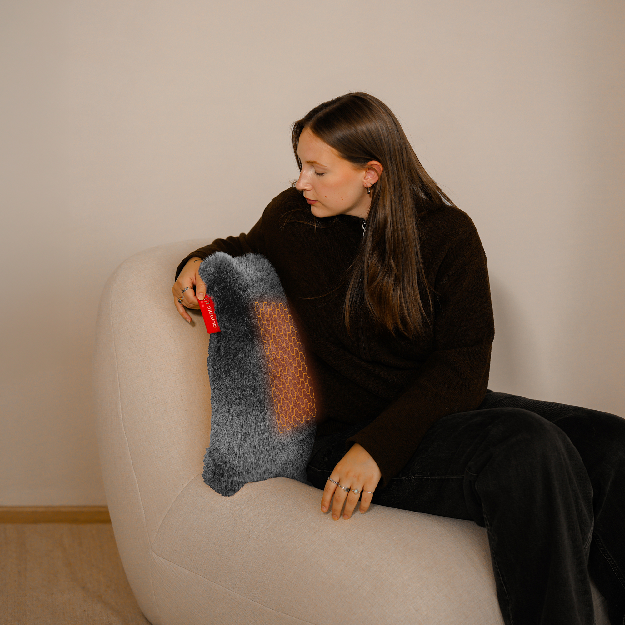 Une femme est assise sur une chaise, regardant un coussin chauffant. Le coussin est gris et a une étiquette rouge. Le fond est un mur beige.