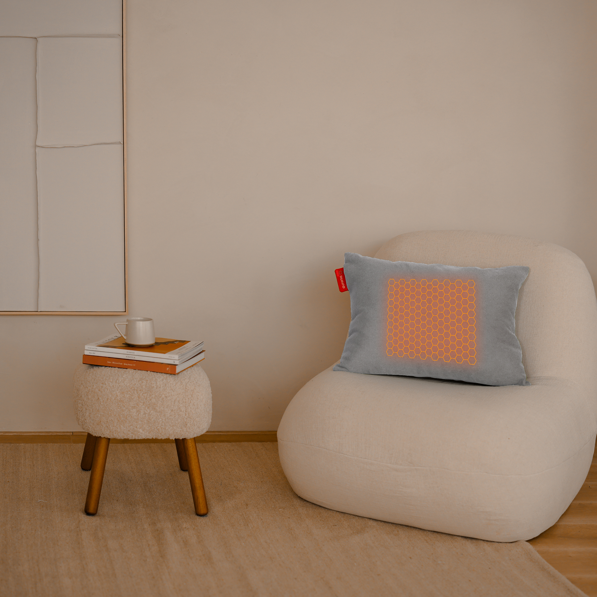 Een grijs kussen met een oranje honingraatpatroon ligt op een witte stoel. Links staat een kleine kruk met boeken en een mok.