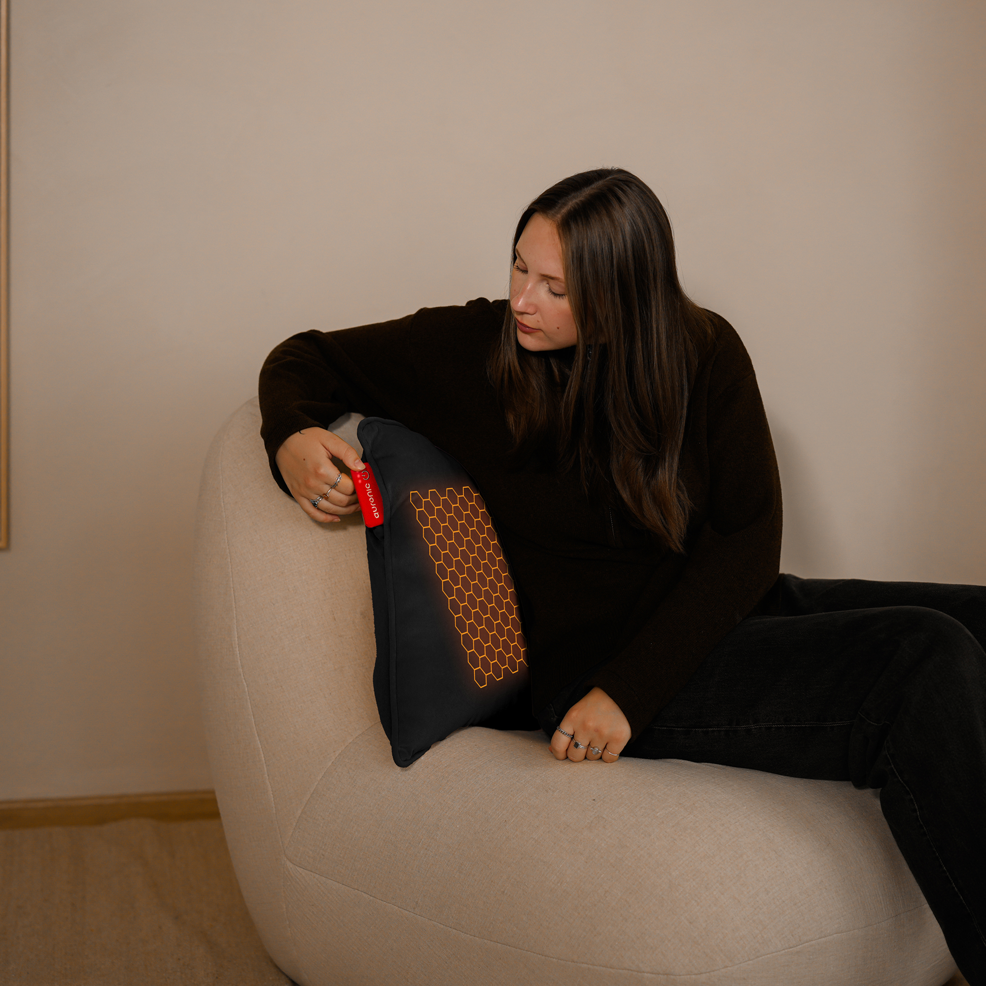 Vrouw die kijkt naar een zwart kussen met een honingraatpatroon, zittend in een beige fauteuil. Ze draagt een donkere trui en een zwarte jeans.