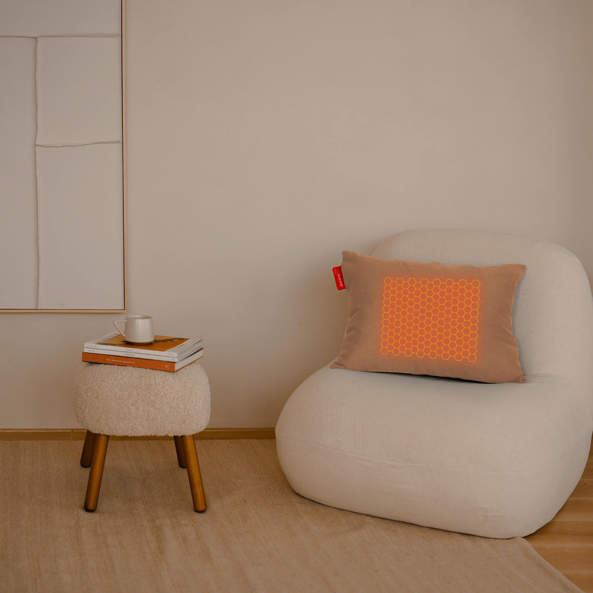 Een beige stoel met een kussen, een bijzettafeltje met boeken en een kopje en een kunstwerk aan de muur.