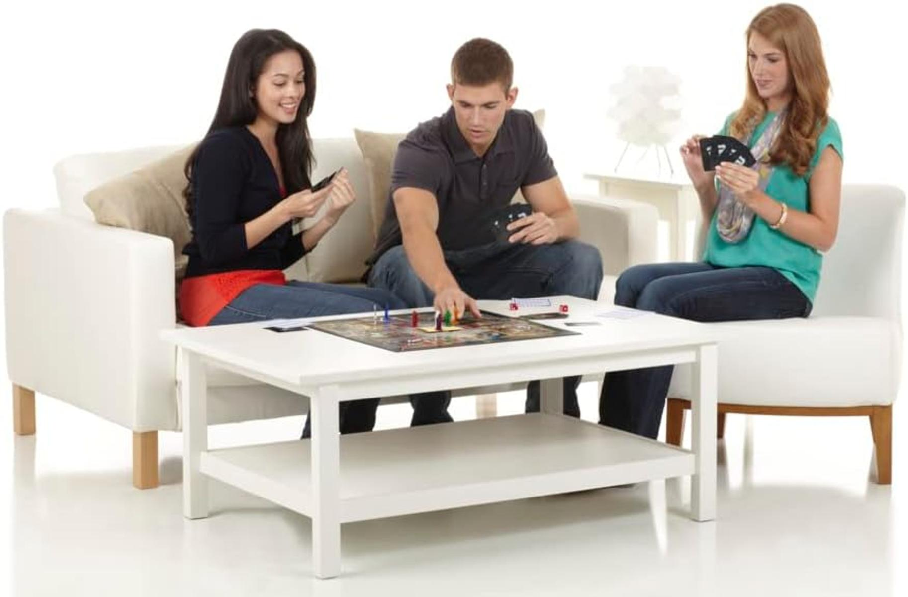 Drei Personen spielen ein Brettspiel auf einem weißen Tisch. Frauen links und rechts halten Karten. Mann in der Mitte bewegt eine Spielfigur.