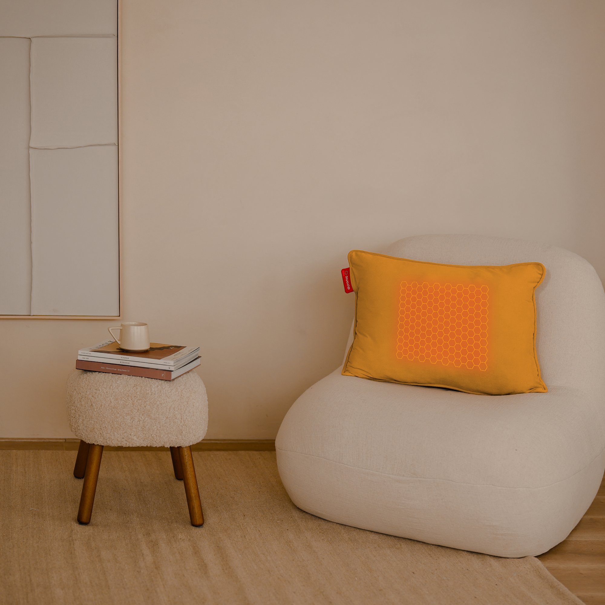 Een kamer met een witte stoel, een oranje kussen met een patroon en een kleine kruk met boeken en een mok.