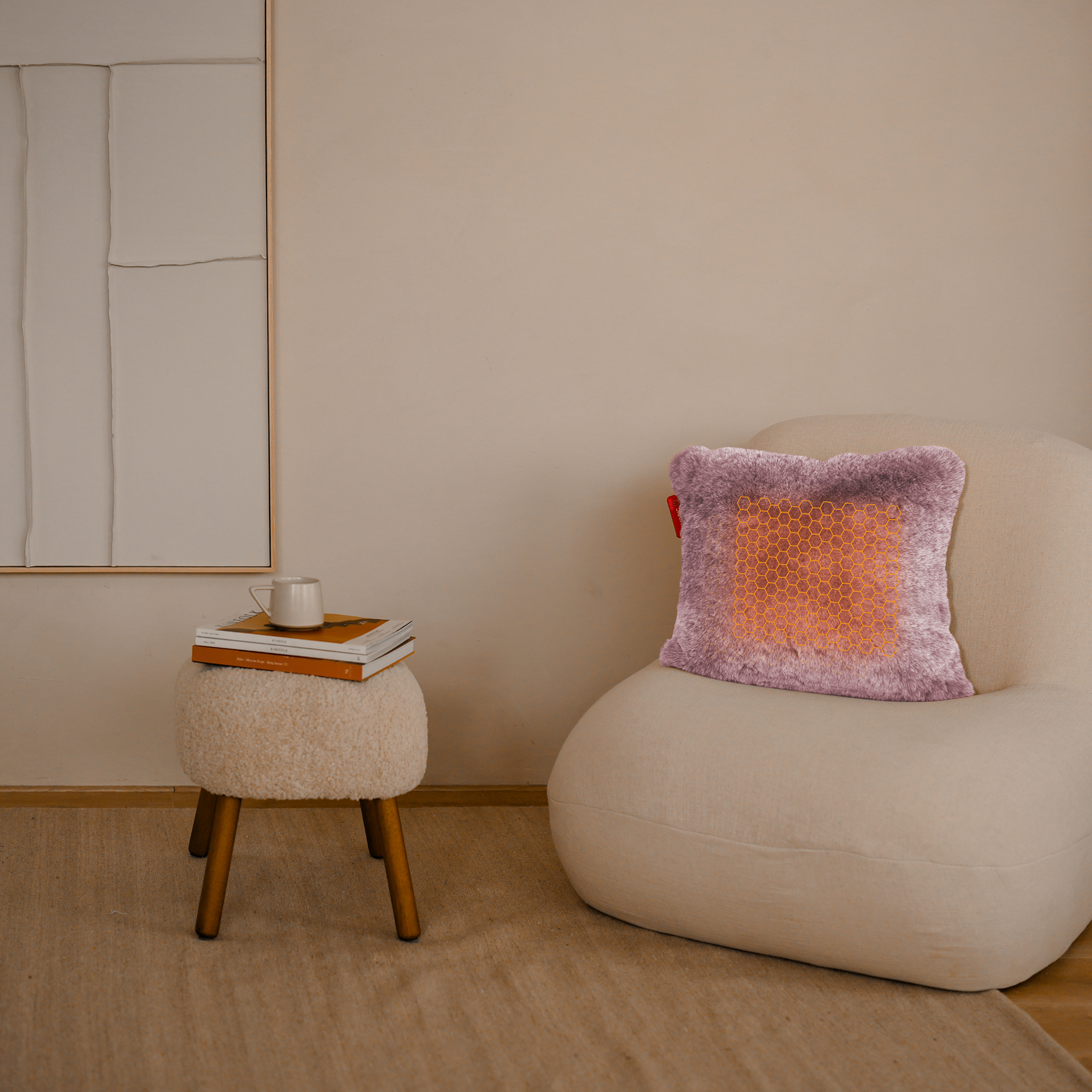 Een kamer met een beige stoel, een zachte kruk met boeken en een kopje, en een paars kussen op de stoel.