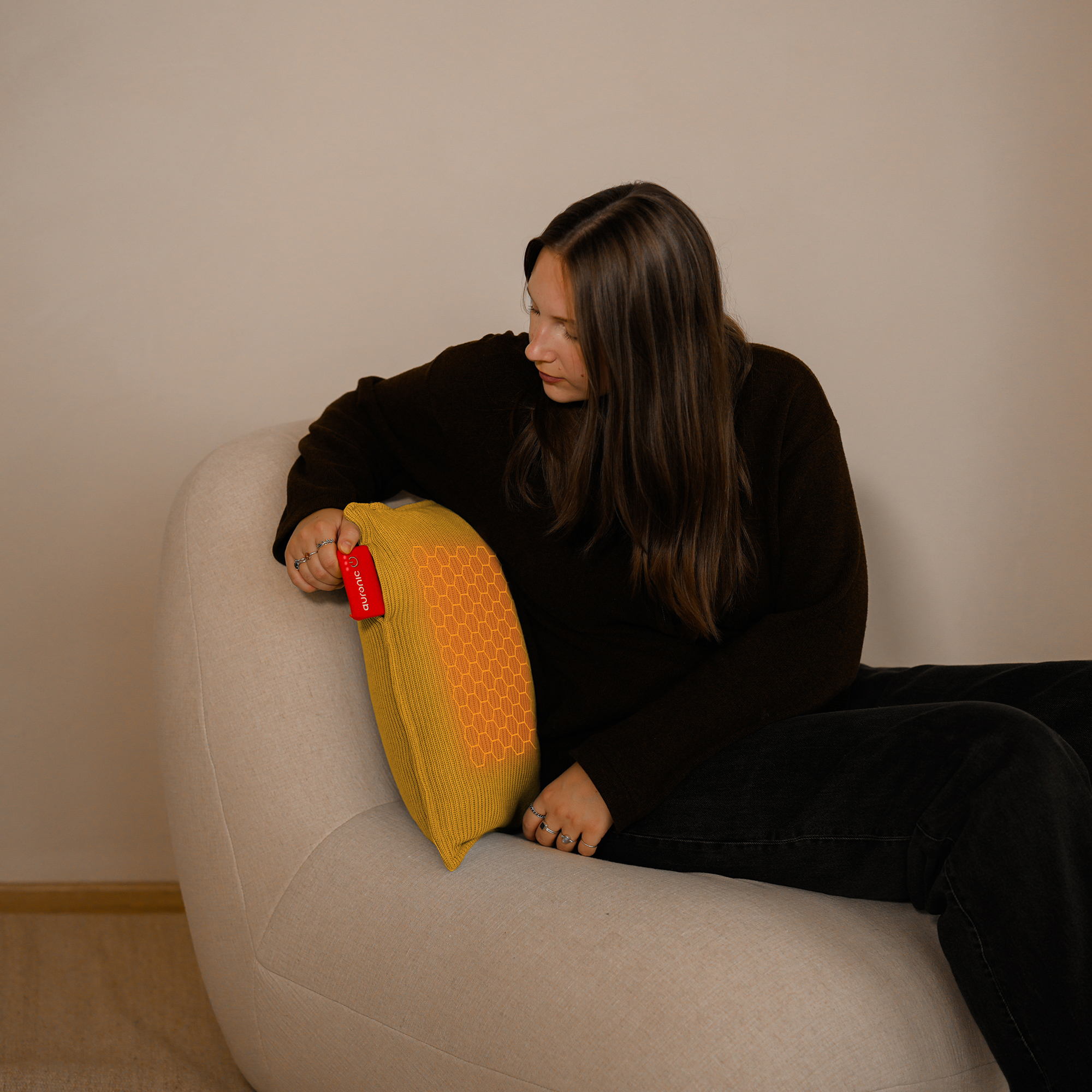 Une femme assise sur une chaise beige tient un oreiller jaune. L'oreiller a une étiquette rouge et un motif en nid d'abeille.