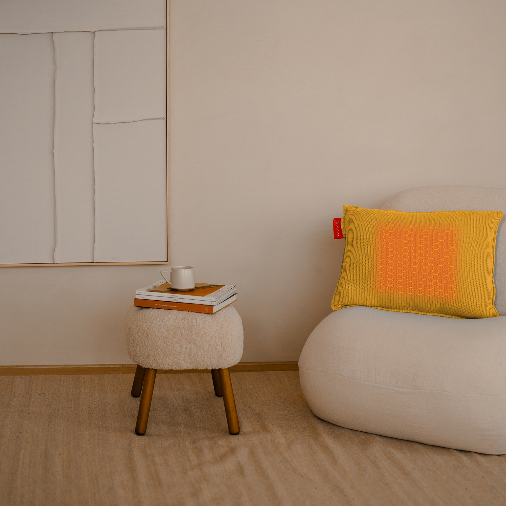 Une pièce avec une chaise blanche, un oreiller jaune, un petit tabouret avec des livres et une tasse. Une image est accrochée au mur.