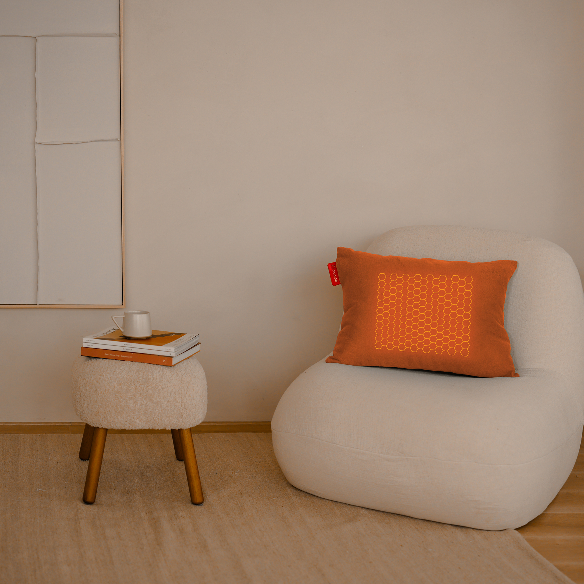 An orange pillow with a honeycomb design on a white chair. A stool with books and a mug is on the left.