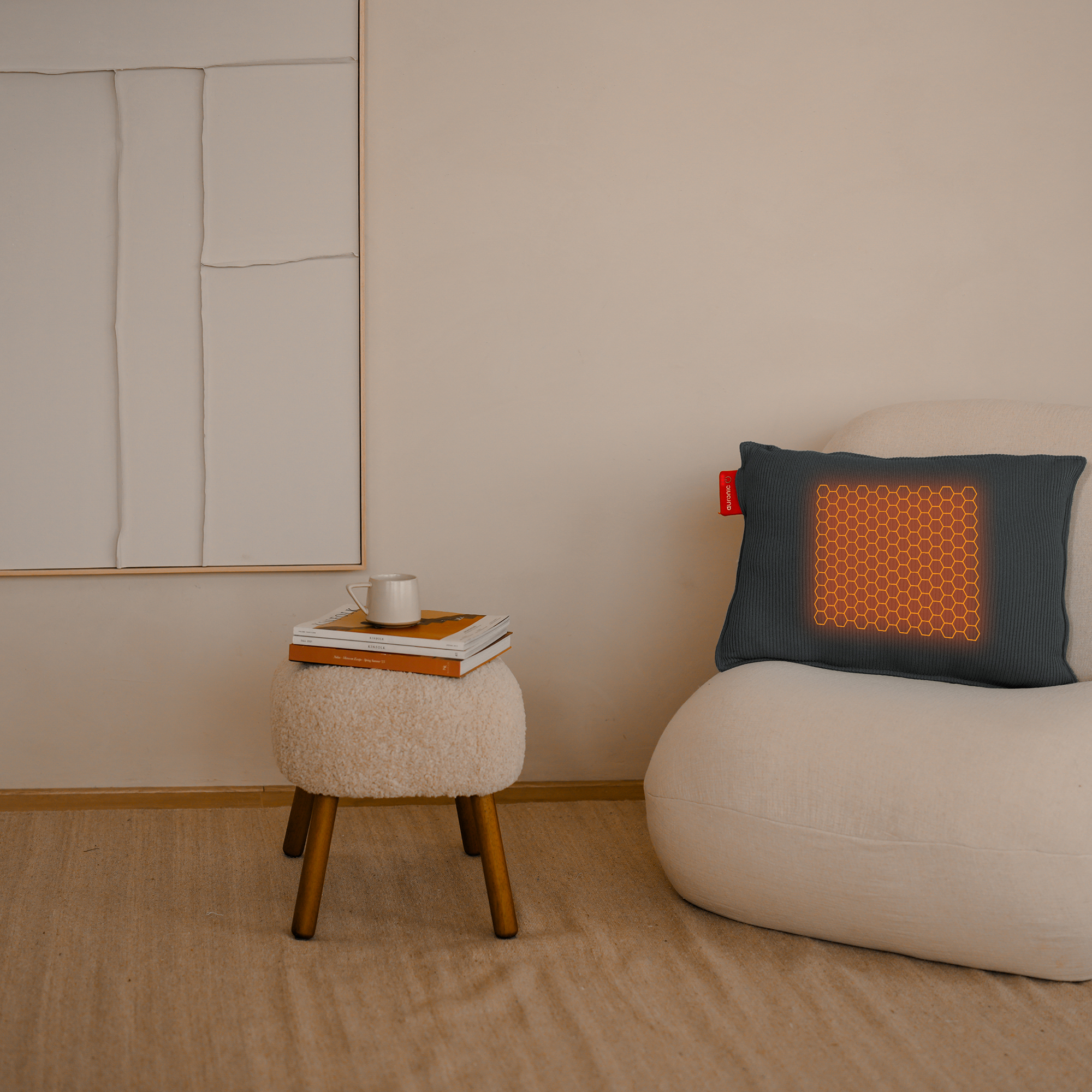Een kamer met een witte stoel, een krukje met boeken en een kopje, een kussen met een gloeiend patroon en een schilderij.