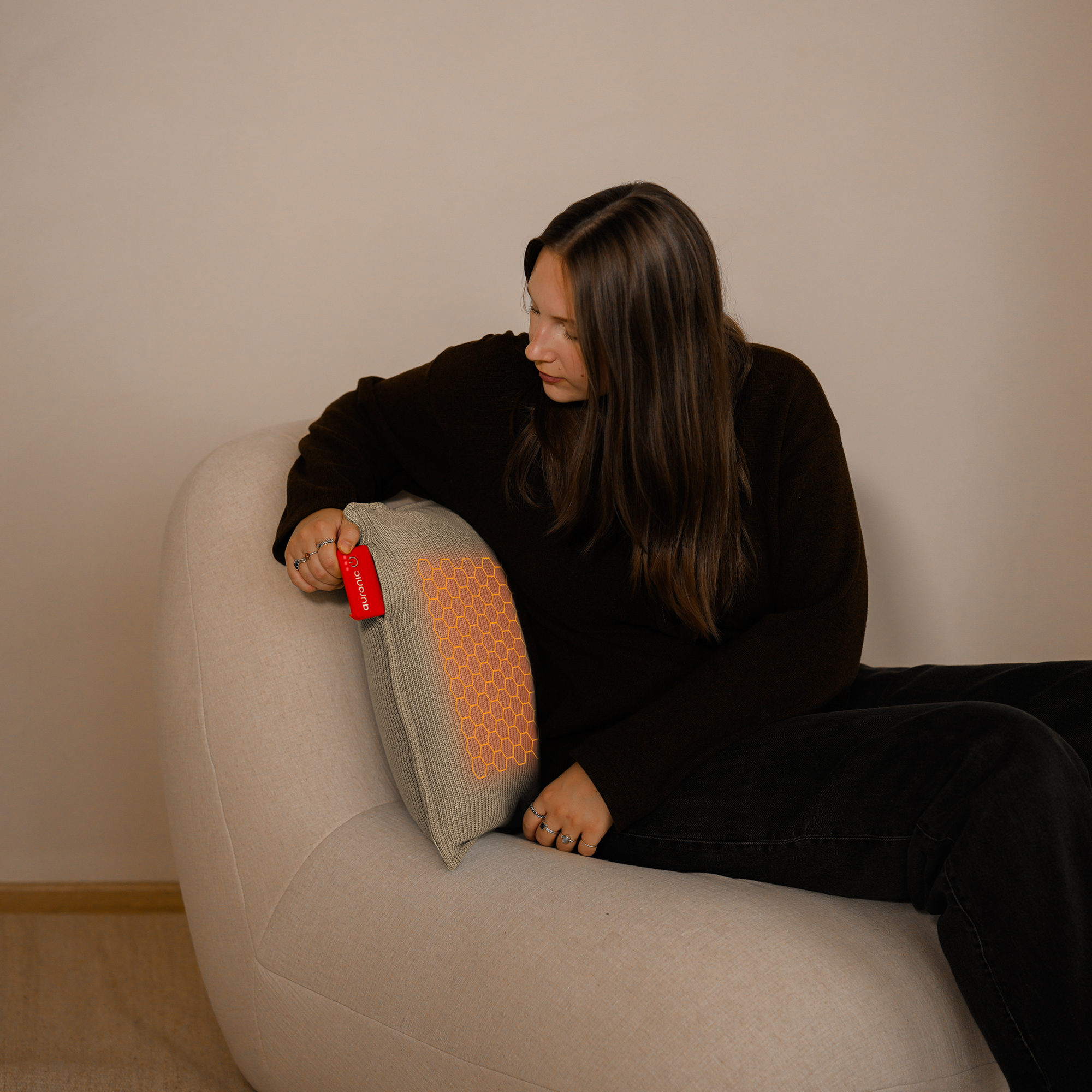 Vrouw zit op een stoel, houdt een rood apparaat vast bij een grijs kussen met een oranje patroon. Ze draagt zwart.