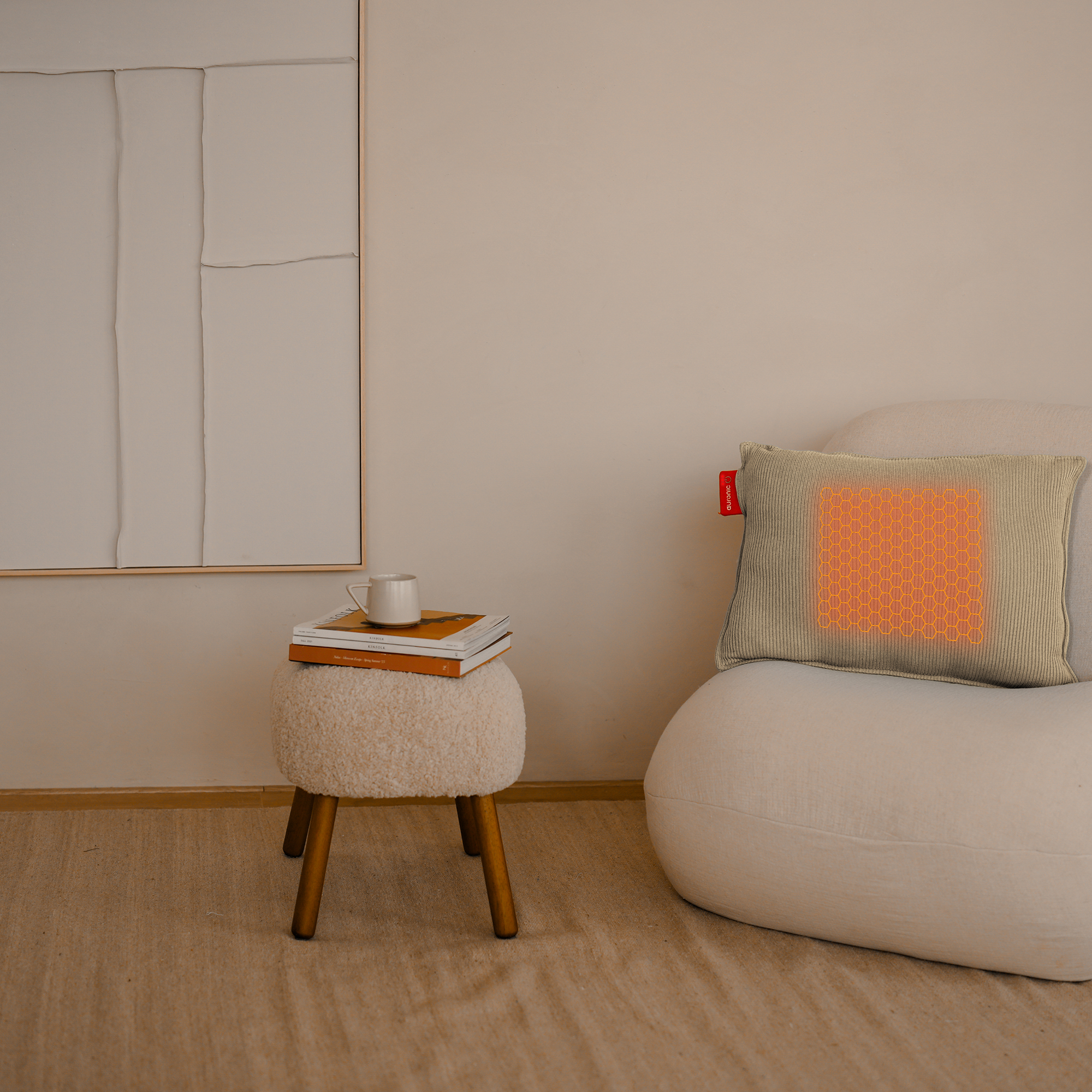 Een beige kussen met oranje warmtepatroon op een witte stoel, krukje met boeken en een mok. Beige muur.