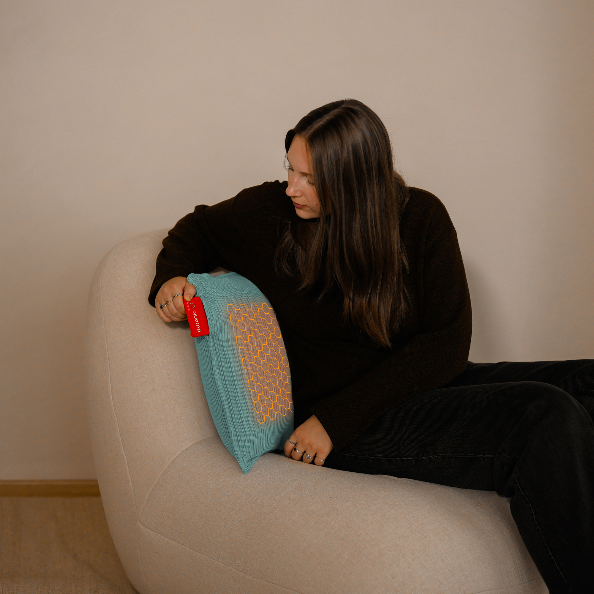 Een vrouw zit op een beige stoel en houdt een blauw kussen vast met een oranje honingraatpatroon. De achtergrond is een effen muur.
