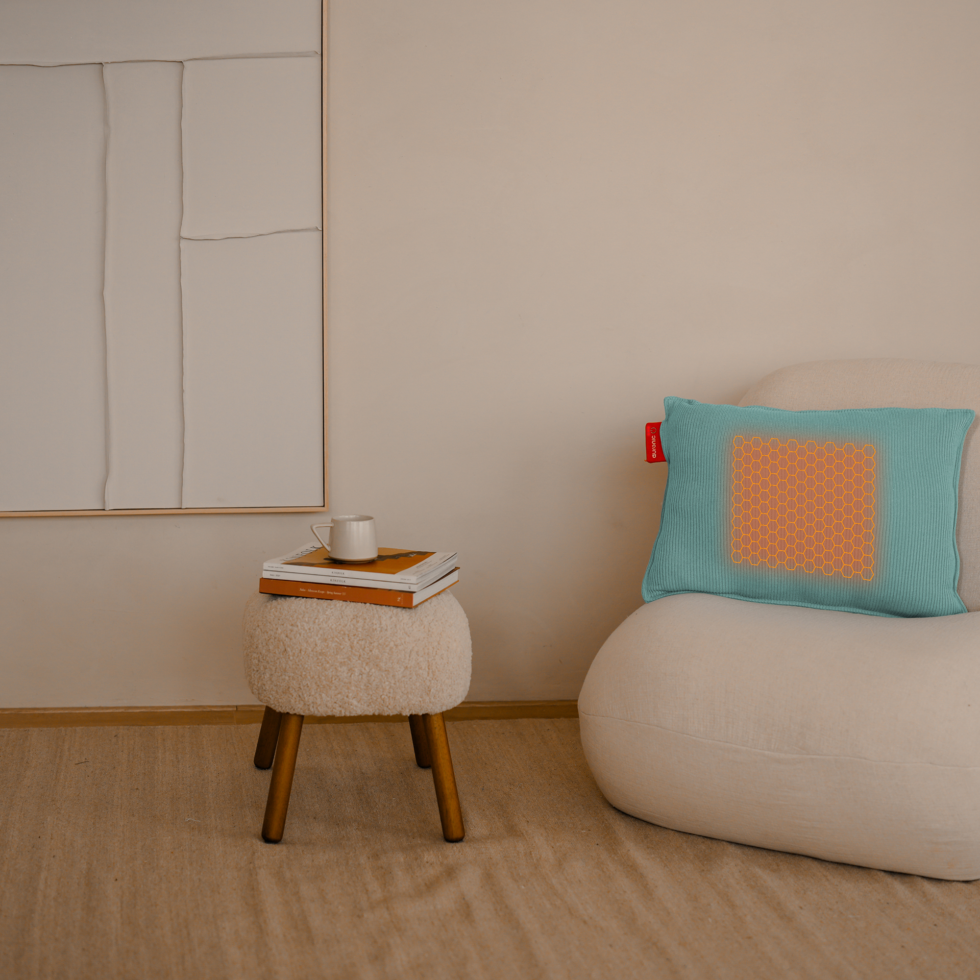Een turquoise kussen met een oranje honingraatontwerp ligt op een crèmekleurige stoel. Een krukje met boeken en een mok is in de buurt.