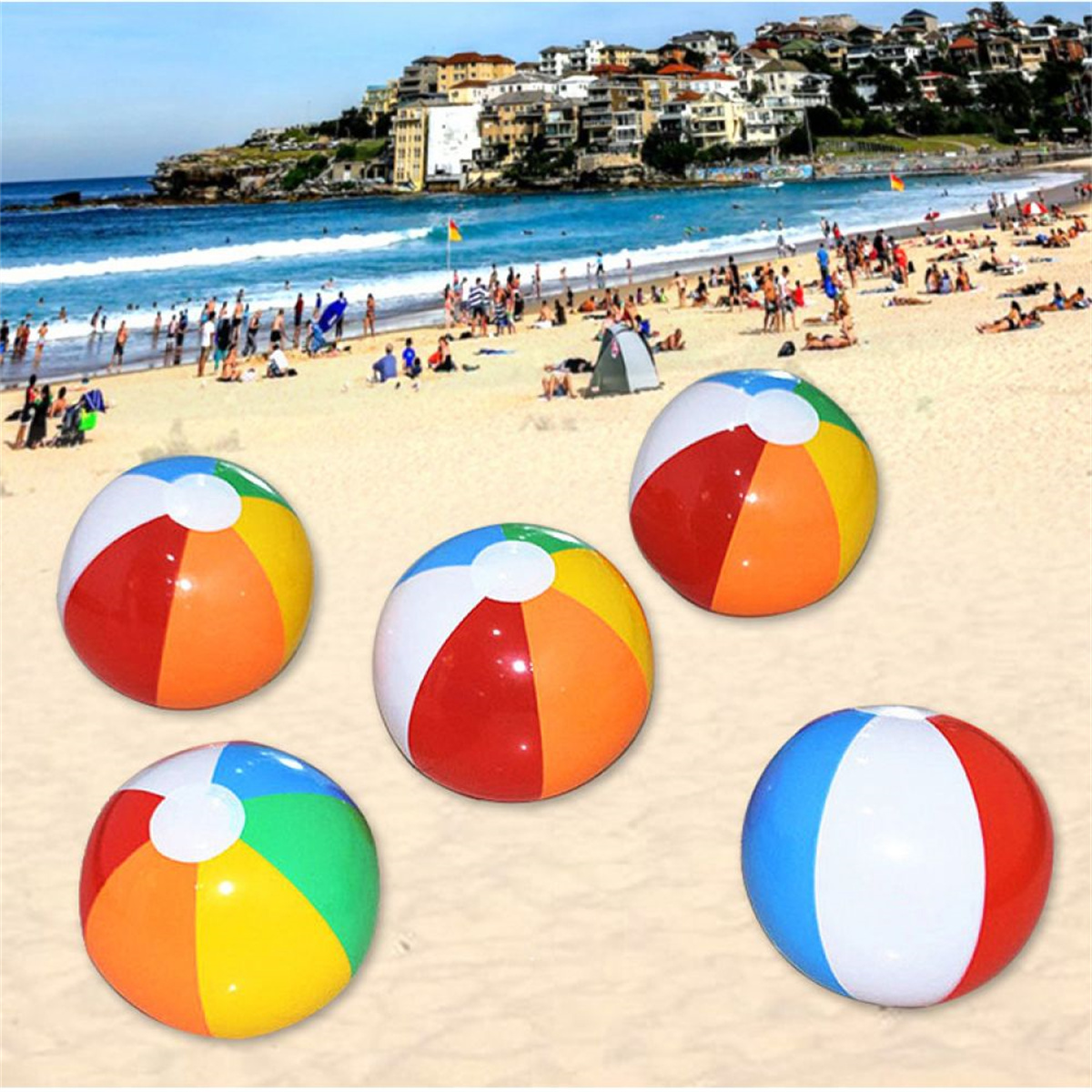 Fünf bunte Strandbälle auf Sand. Strand mit Leuten, Gebäuden im Hintergrund und blauem Wasser.