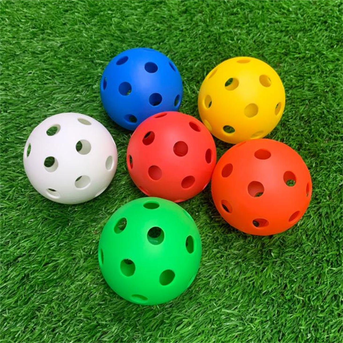 Sechs bunte Plastikbälle mit Löchern auf grünem Gras. Farben sind Weiß, Blau, Gelb, Rot, Orange und Grün.
