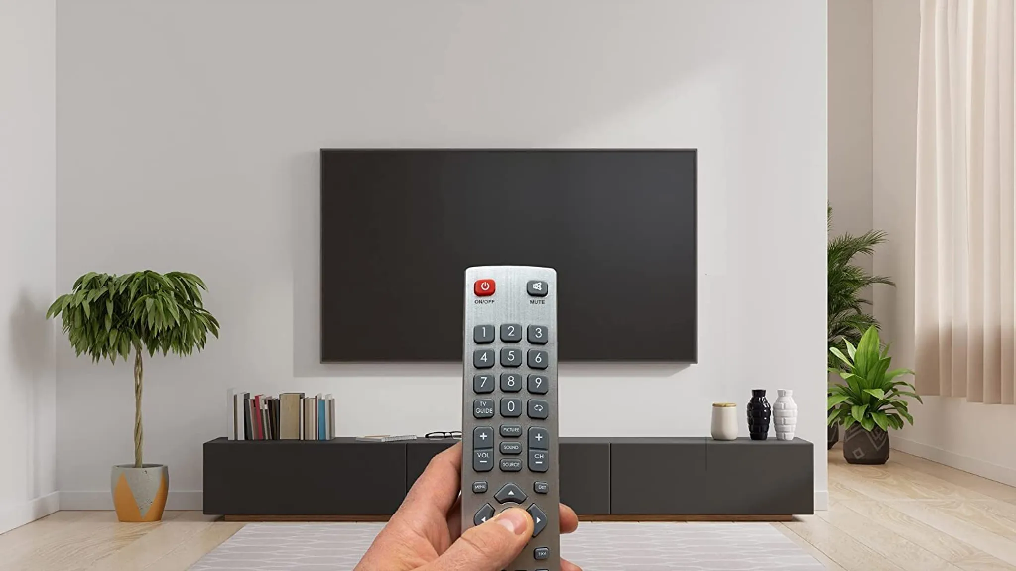 Hand hält TV-Fernbedienung, die auf einen großen schwarzen Bildschirm zeigt. Ein kleiner Baum und Dekorationsartikel sind sichtbar.