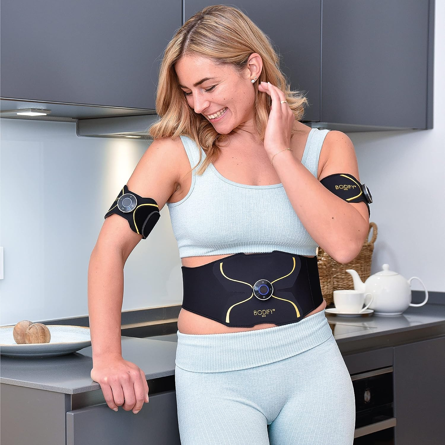 Frau mit Fitnessgeräten lächelt in einer Küche. Schwarze und gelbe Geräte an Armen und Taille. Hellblaues Outfit.