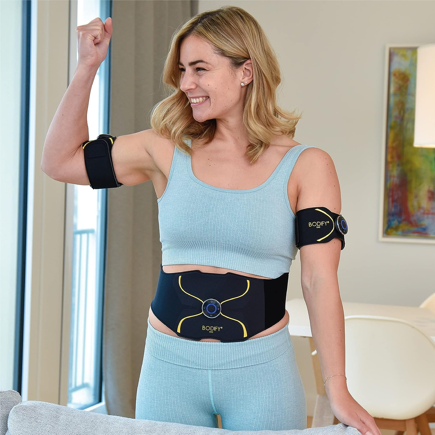 Eine Frau lächelt, trägt blaue Kleidung und Fitnessgeräte. Sie hebt den Arm. Hintergrund: Fenster, Gemälde und Möbel.