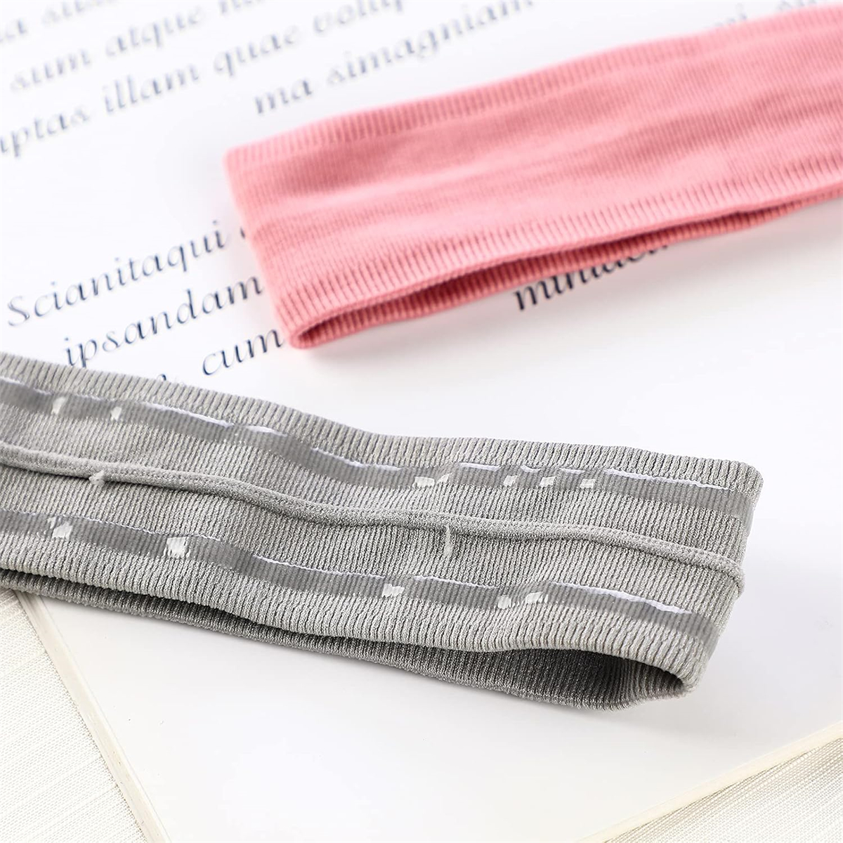 Zwei Stirnbänder auf weißem Untergrund mit Text. Eines rosa, eines grau mit weißen Linien. Hintergrund hat gedruckten Text.