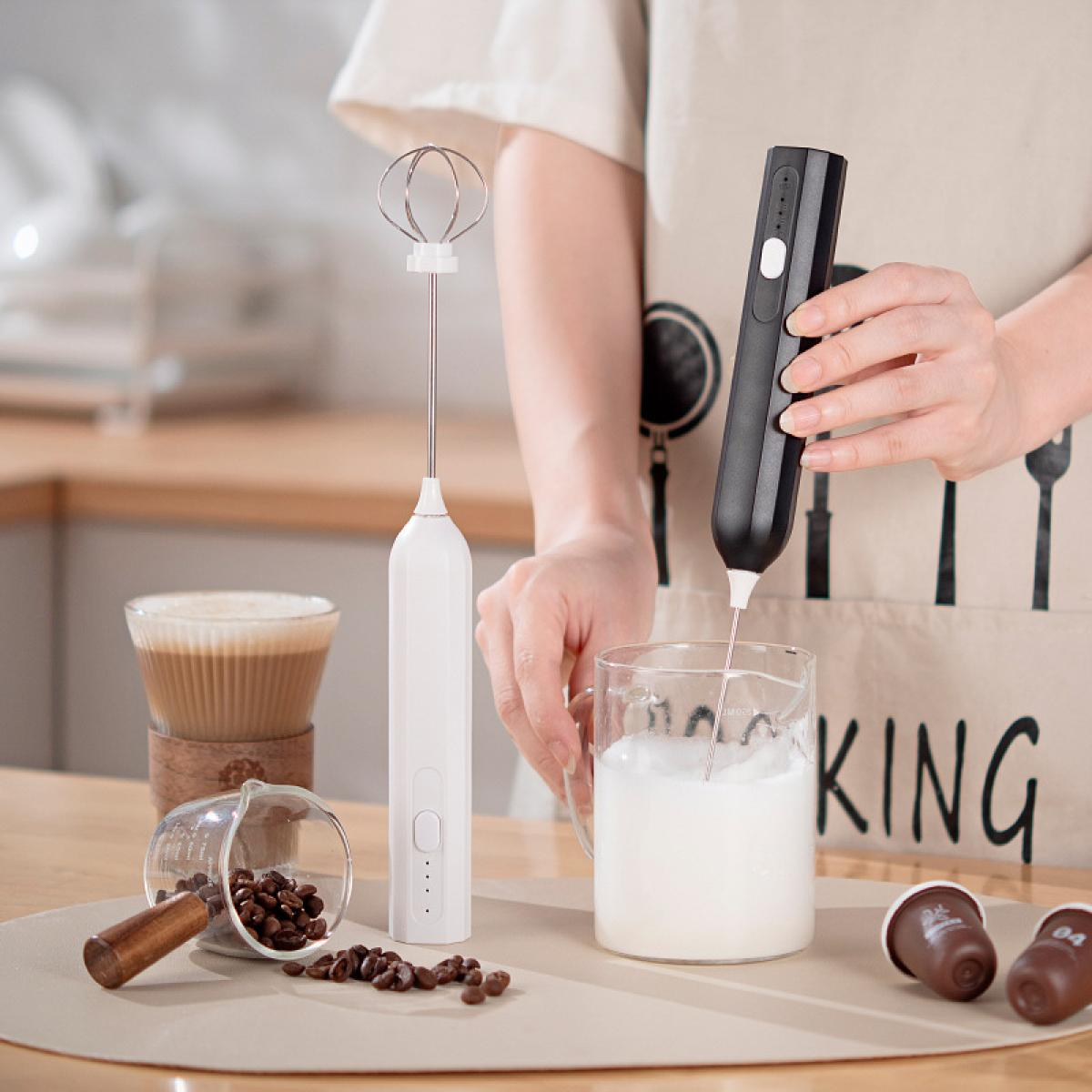 Eine Person schäumt Milch mit einem schwarzen Aufschäumer auf. Kaffeebohnen, ein weißer Aufschäumer und Kaffee stehen auf dem Tisch.