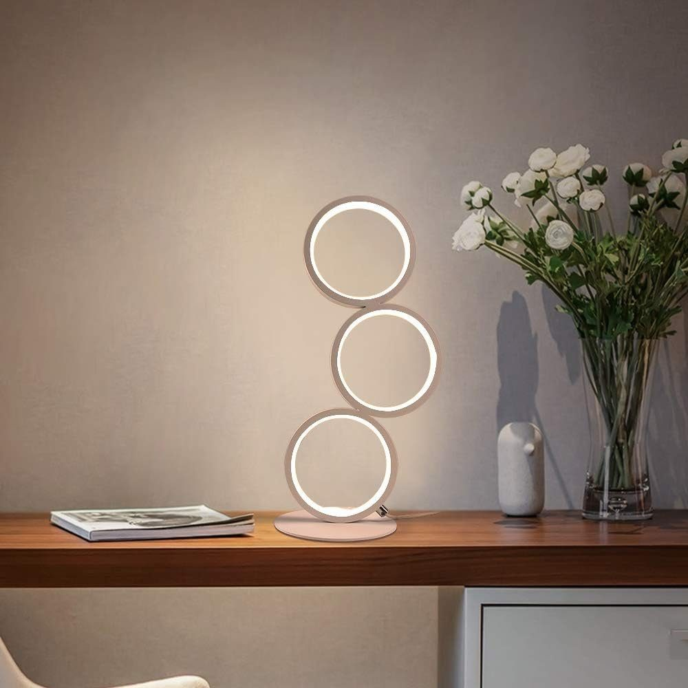 Una lampada moderna con tre lampade circolari si trova su una scrivania di legno. Fiori bianchi in un vaso a destra.