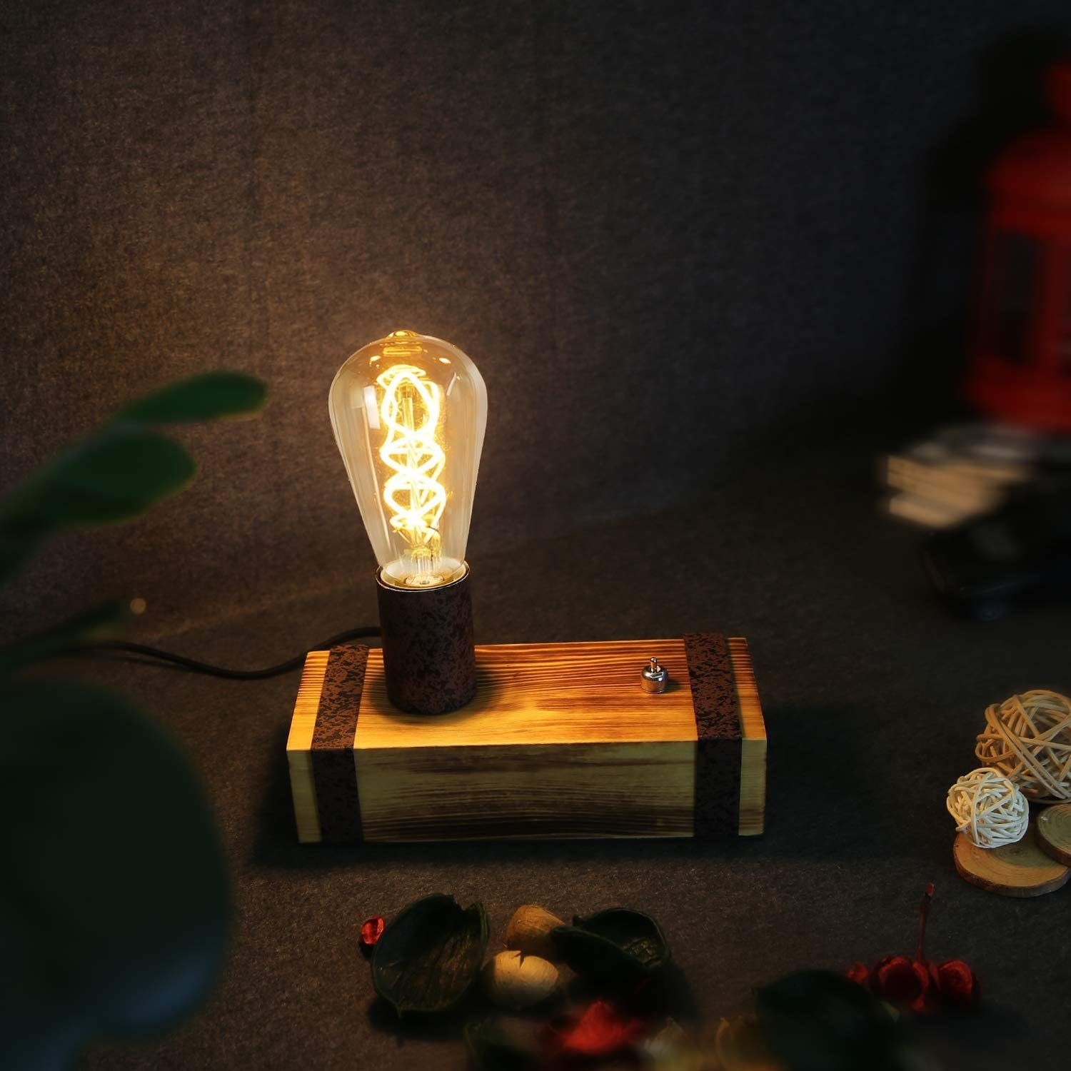 Una lampada con lampadina Edison accesa su una base di legno. Fili, oggetti decorativi e foglie sfocate sullo sfondo.