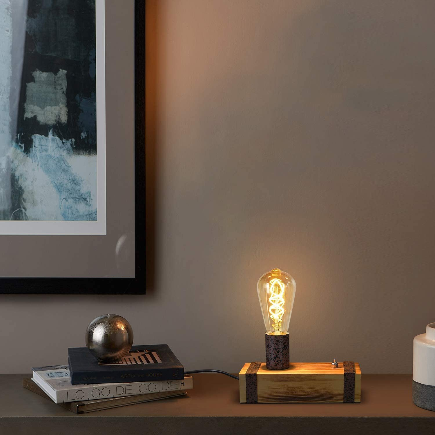 Una lampada con lampadina accesa con base in legno è su un tavolo. Sul tavolo ci sono anche libri e una sfera d'argento. Un dipinto è appeso al muro.