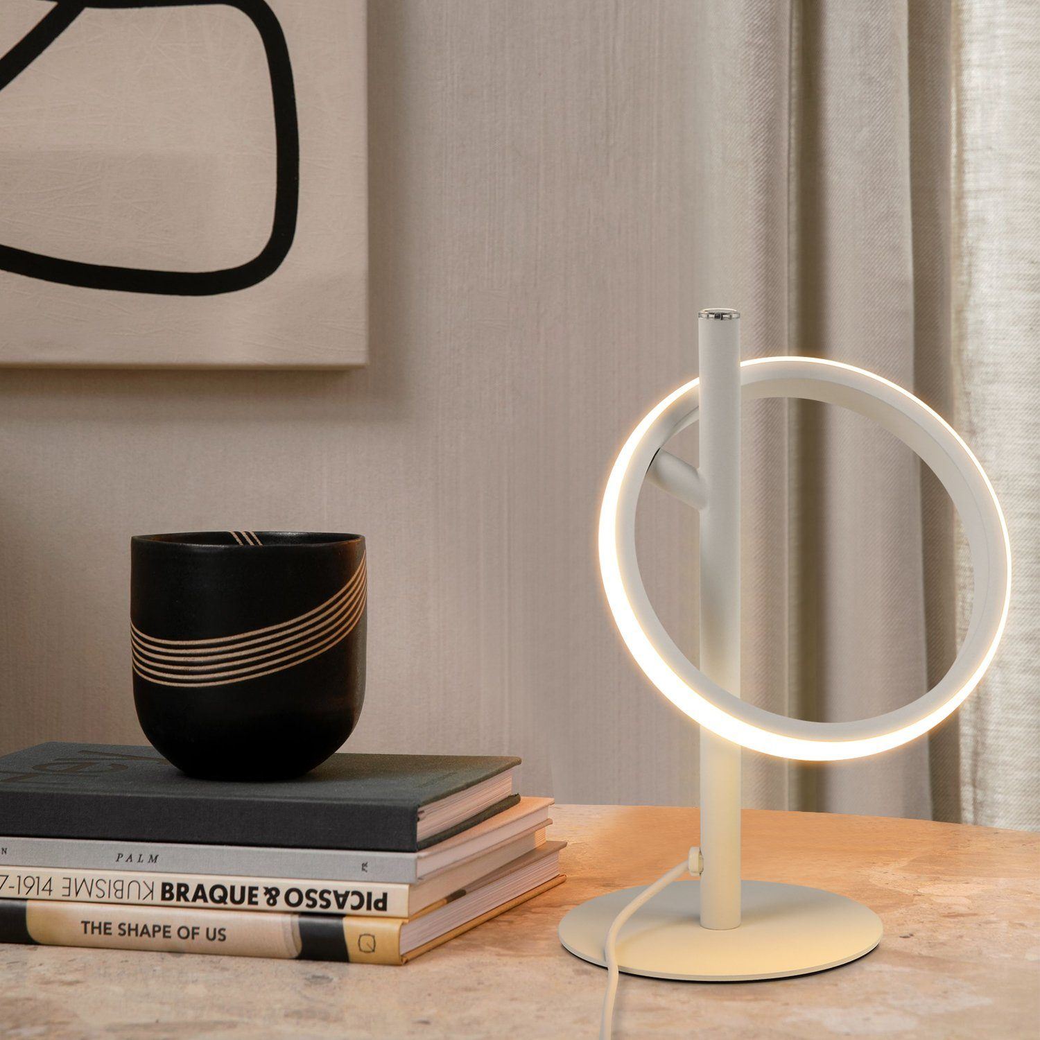 Una lámpara moderna con luz circular, libros y una taza negra sobre una mesa de madera. Fondo neutro con cortina beige.