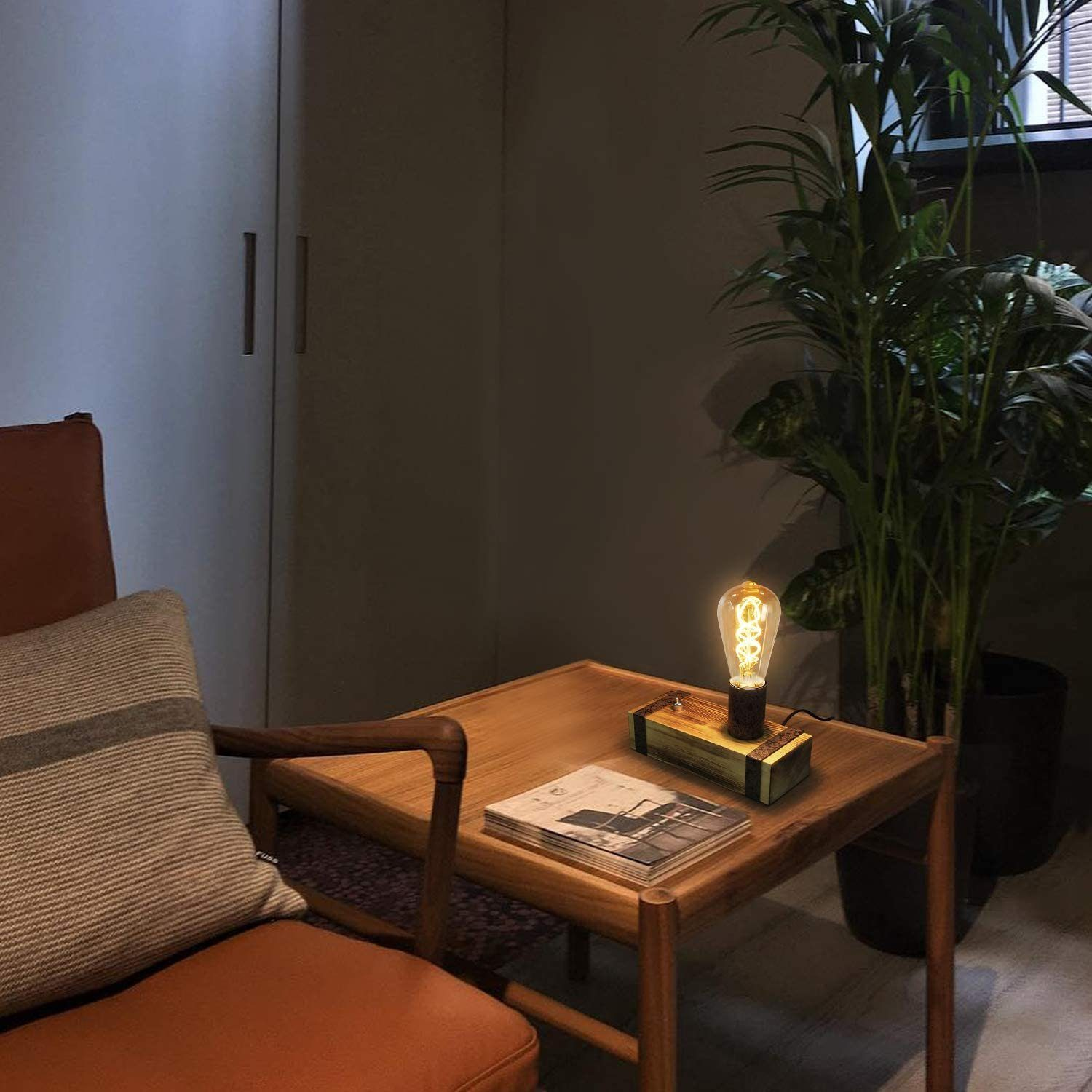 Una lampada con lampadina accesa su un tavolo di legno con una rivista. Una pianta sullo sfondo e una sedia a sinistra.