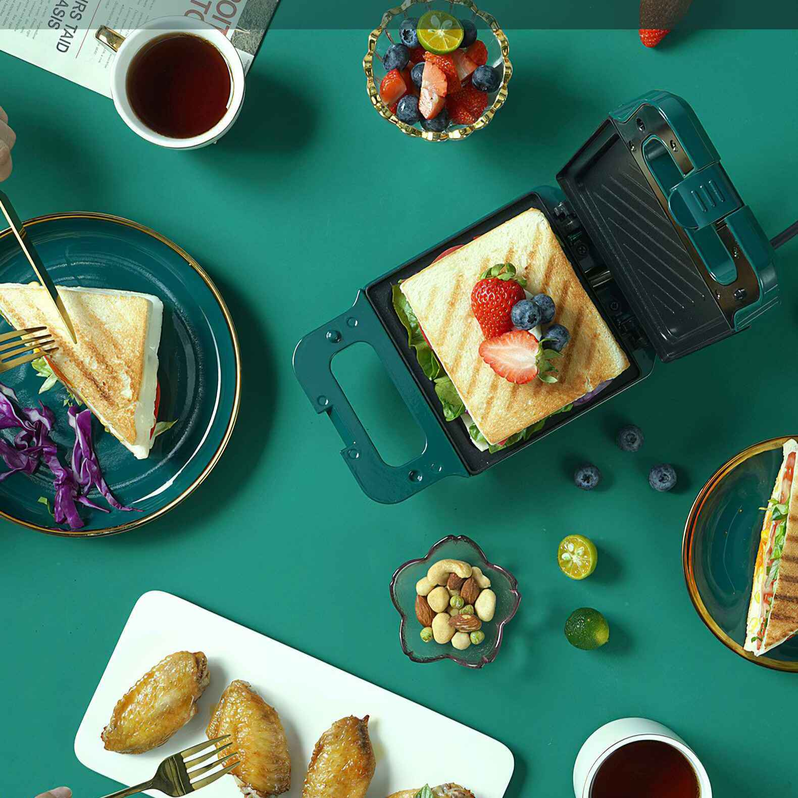 Eine grüne Tischdecke mit Sandwichmaker, Sandwiches, Früchten, Nüssen, Flügeln und Getränken.