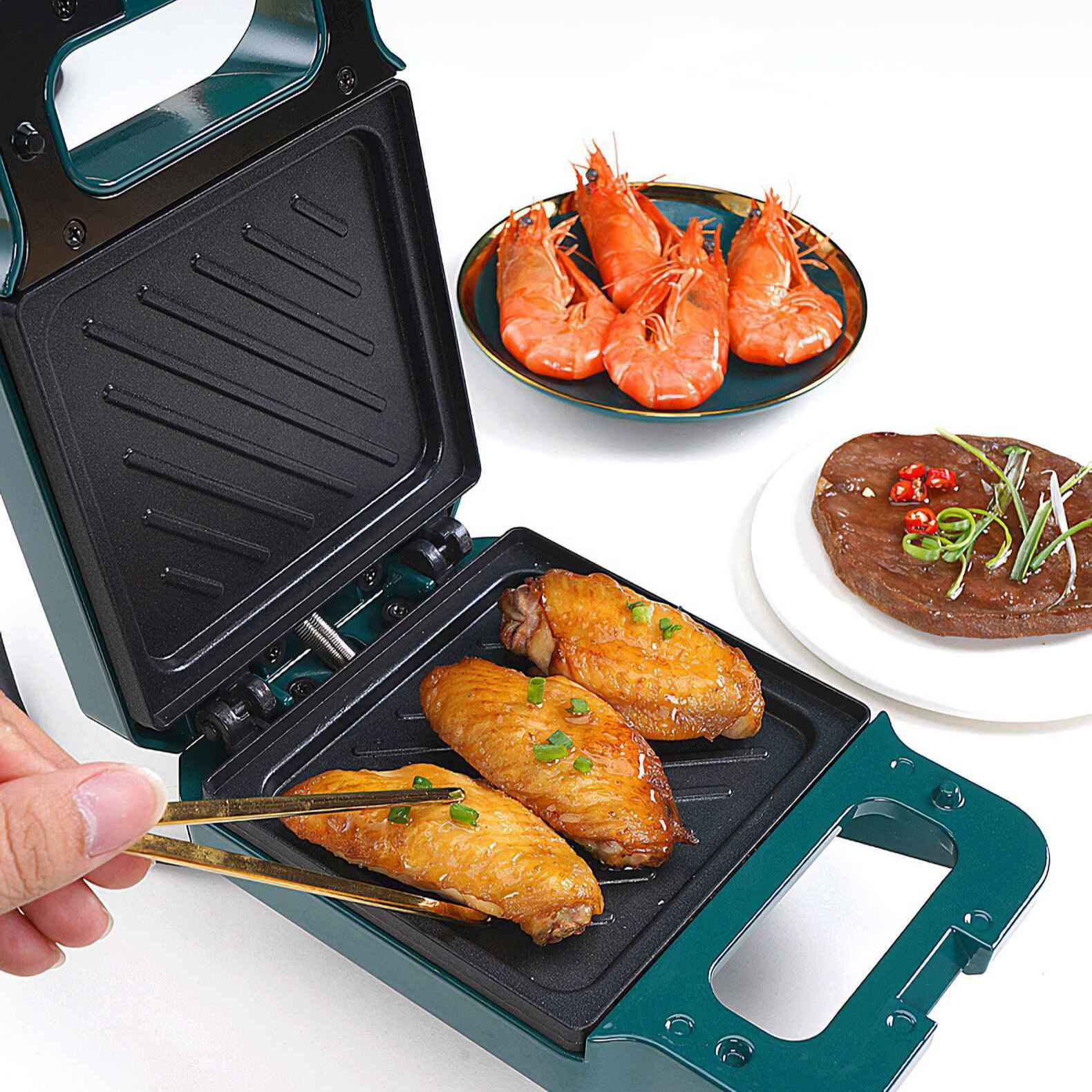 Ein Sandwichgrill mit Hähnchenflügeln, Garnelen und Steak. Eine Hand hält eine Zange. Der Hintergrund ist weiß.