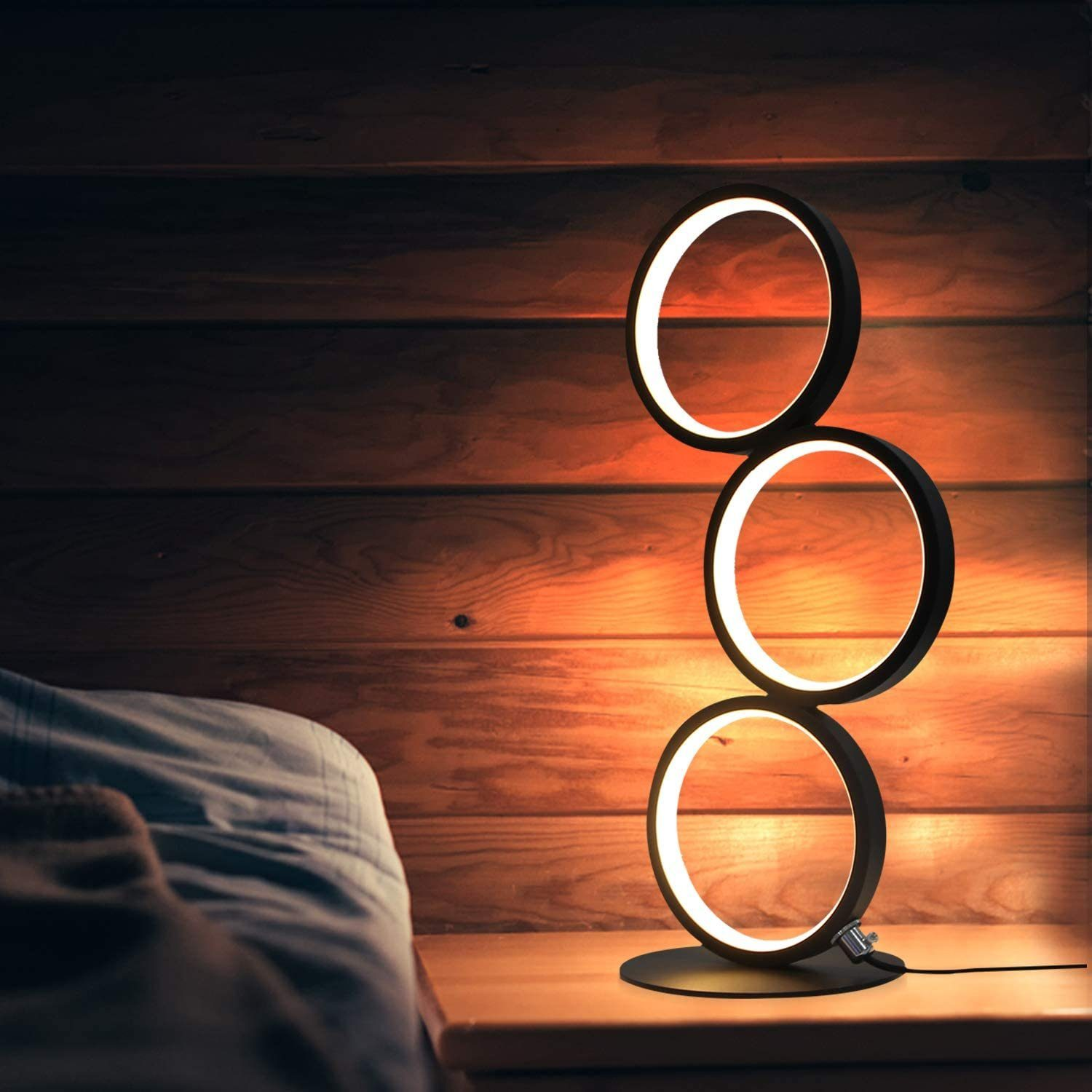 Una moderna lampada nera con tre anelli illuminati su un comodino. Un letto in primo piano, con assi di legno sullo sfondo.